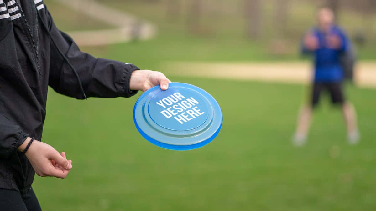 Disc Golf 桌布 壁紙 — 一個穿著黑色外套的人拿著一個有文字的藍色飛盤，準備在草地上拋出，背景有另一個模糊的玩家。