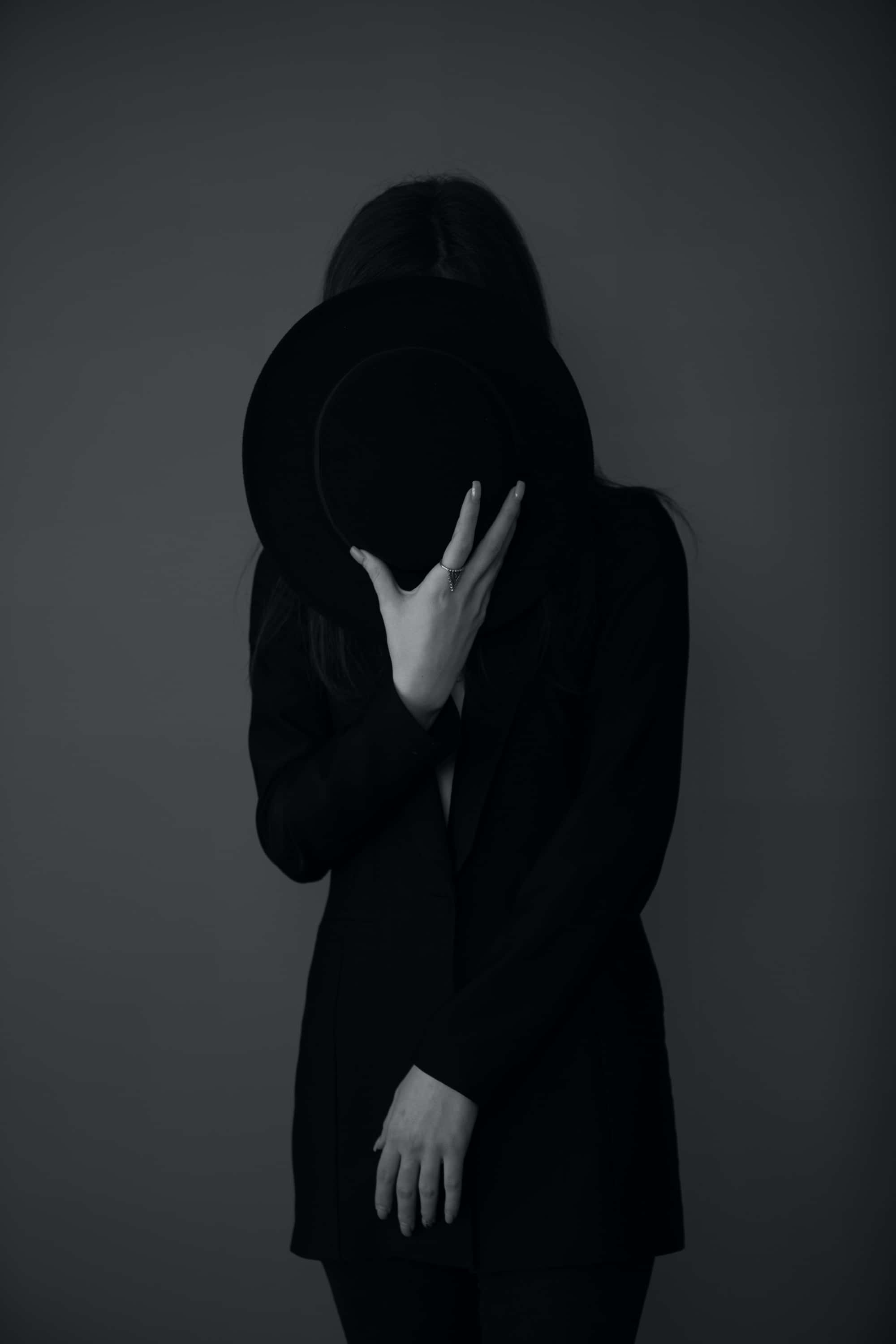 Woman 桌布 壁紙 — 一個穿著黑色外套的人用寬邊帽遮住臉，背景是柔和的灰色。