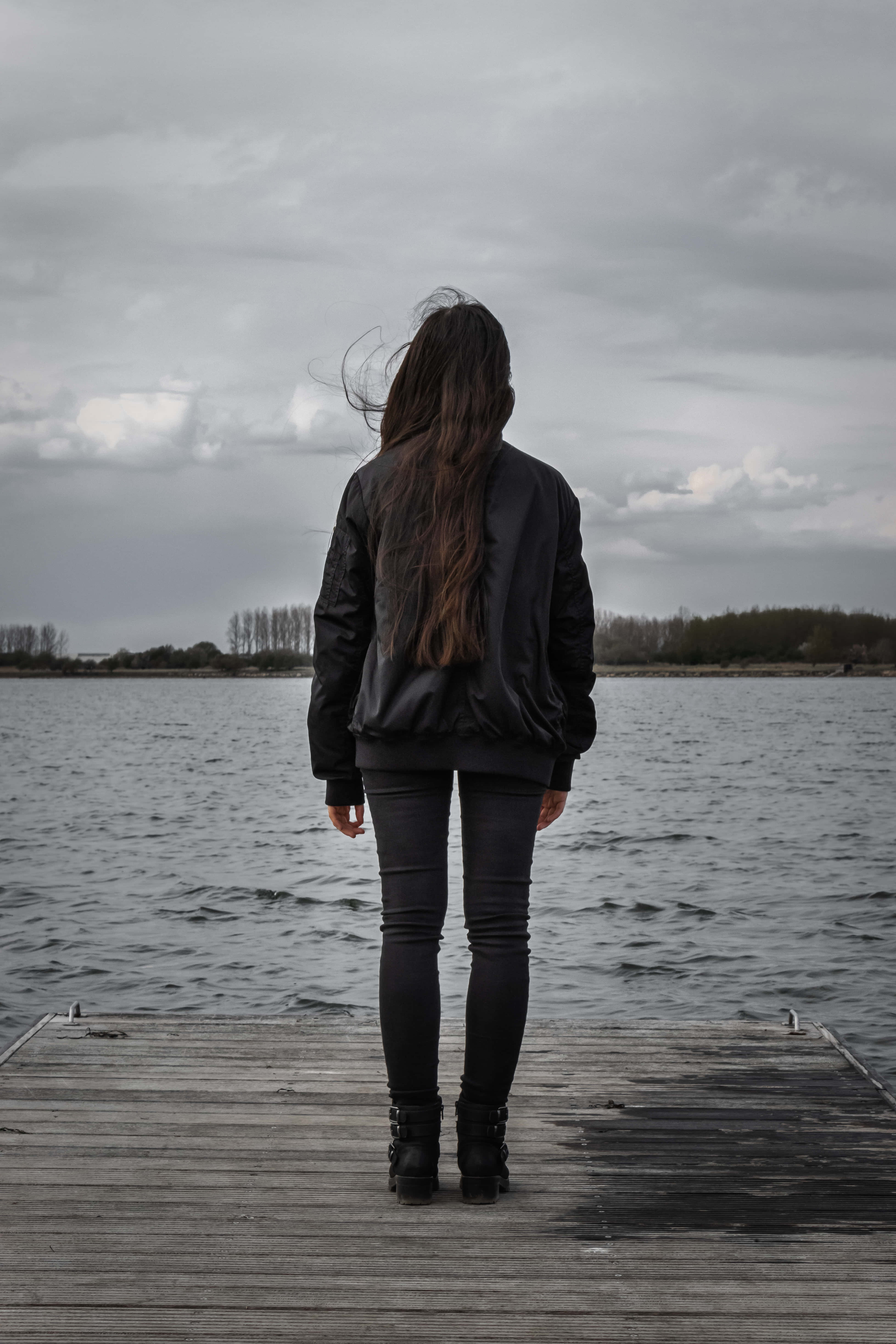 Alone 桌布 壁紙 — 一個穿著黑色外套的孤獨女人站在一個風化的木碼頭上，面對著陰天下一片廣闊的灰色湖泊。