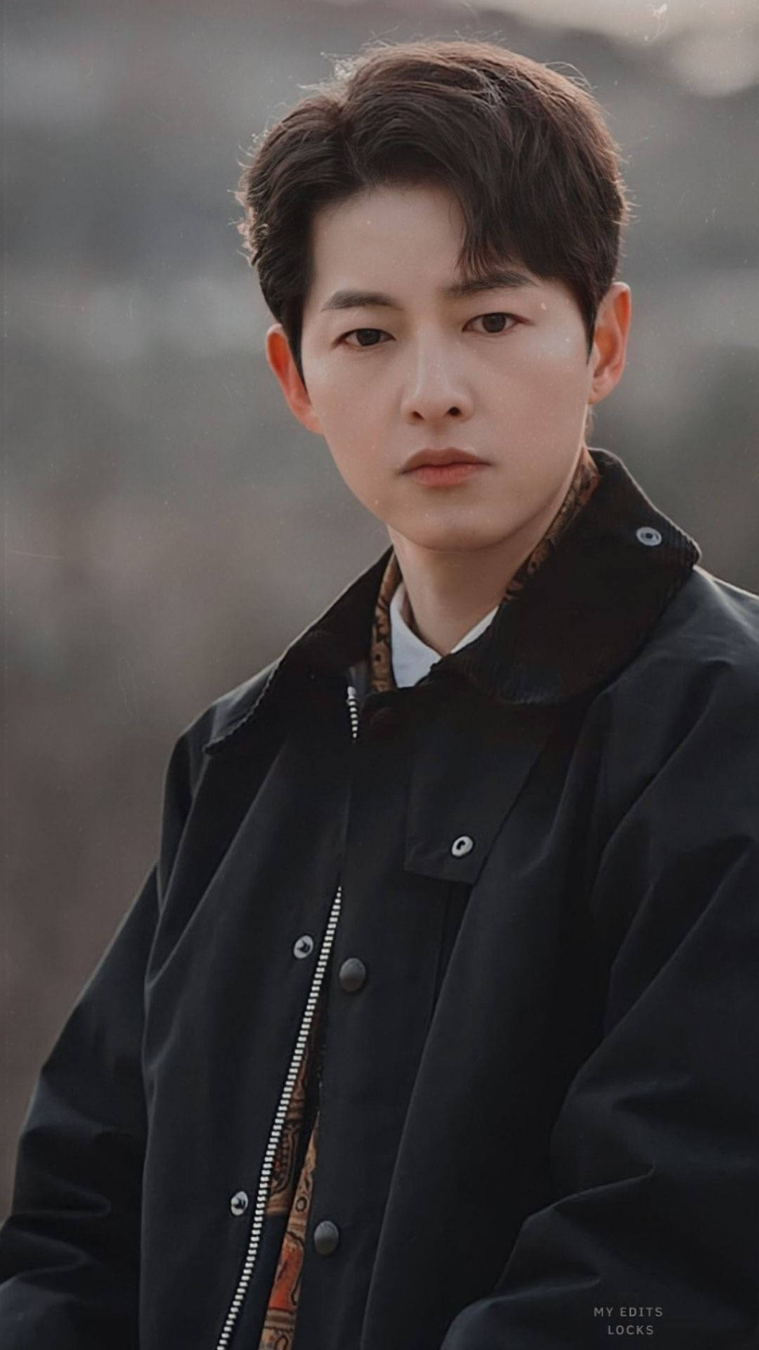 Vincenzo 桌布 壁紙 — 一個穿著黑色外套的年輕男子，在柔和模糊的戶外背景下平靜地凝視著鏡頭。