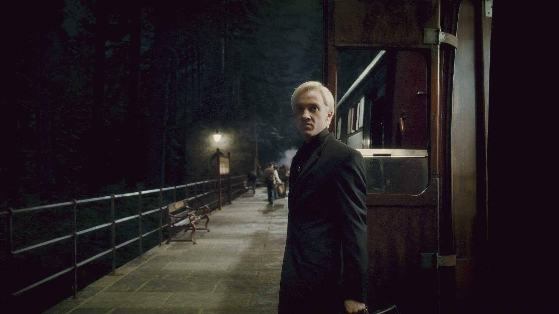 draco 桌布 壁紙 — 一個穿著黑色外套的年輕男子夜晚站在燈光昏暗的火車站月台上，周圍是黑暗的樹木和霧氣。