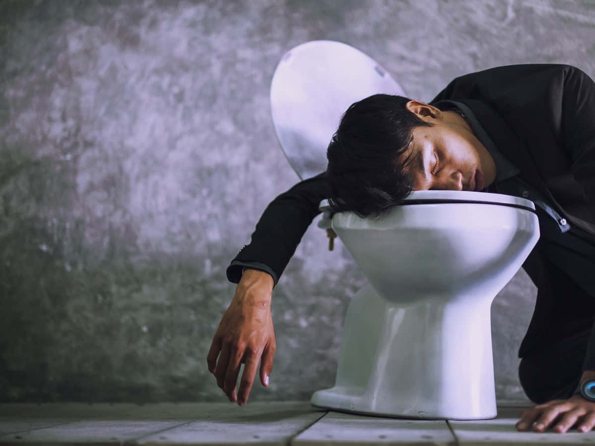 醉意朦朧 桌布 壁紙 — 一個穿著黑色外套的男人在一個牆壁破裂、地板鋪瓷磚的骯髒浴室裡對著馬桶嘔吐。