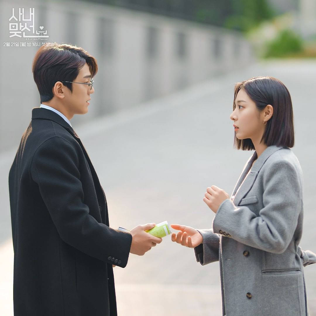 Proposal 桌布 壁紙 — 一個穿著黑色外套的男人在鋪砌的戶外小徑上向一個穿著灰色外套的女人遞上一杯綠色飲料。