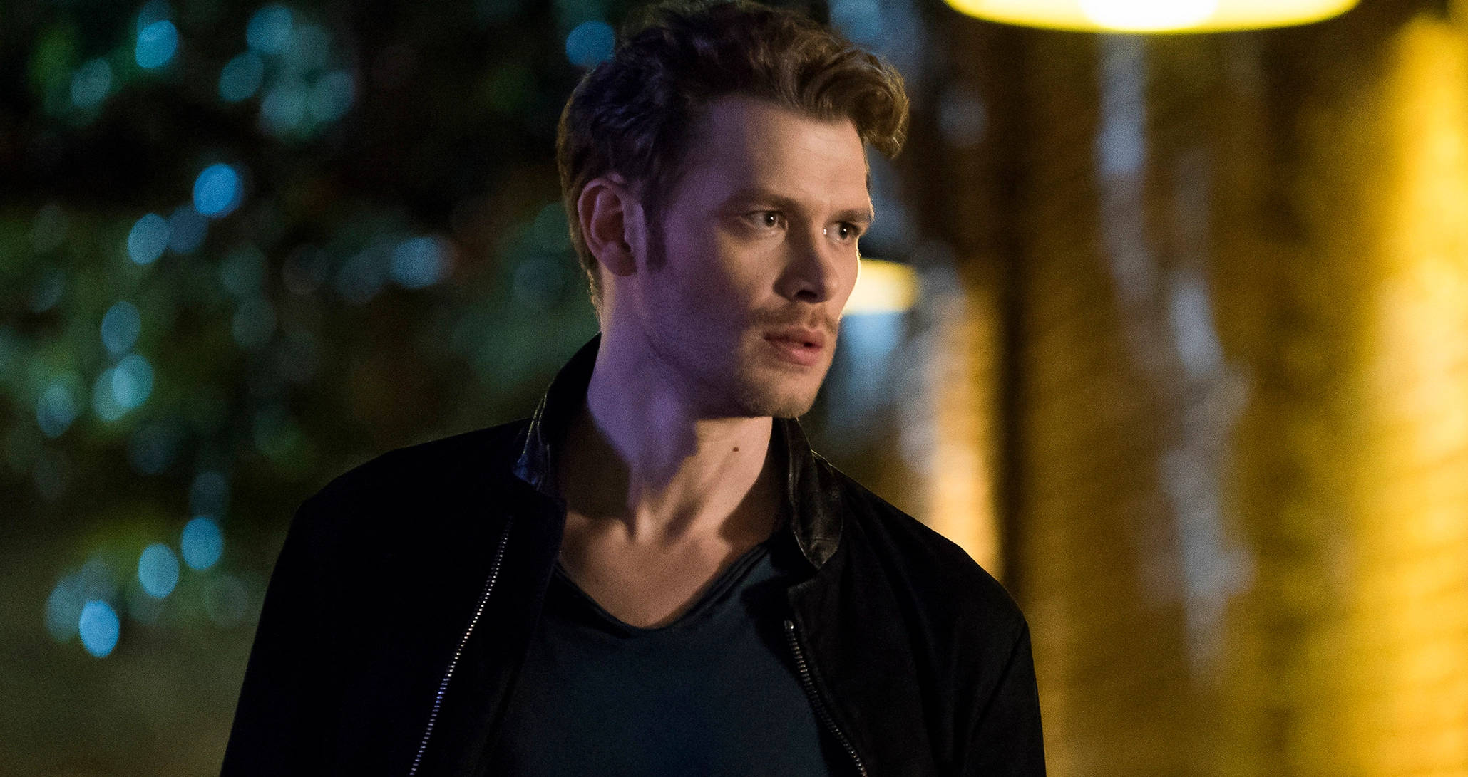 Vampire Diaries 桌布 壁紙 — 一個穿著黑色外套的男人站在昏暗的夜晚，背景有溫暖的黃色燈光和冷藍色散景。