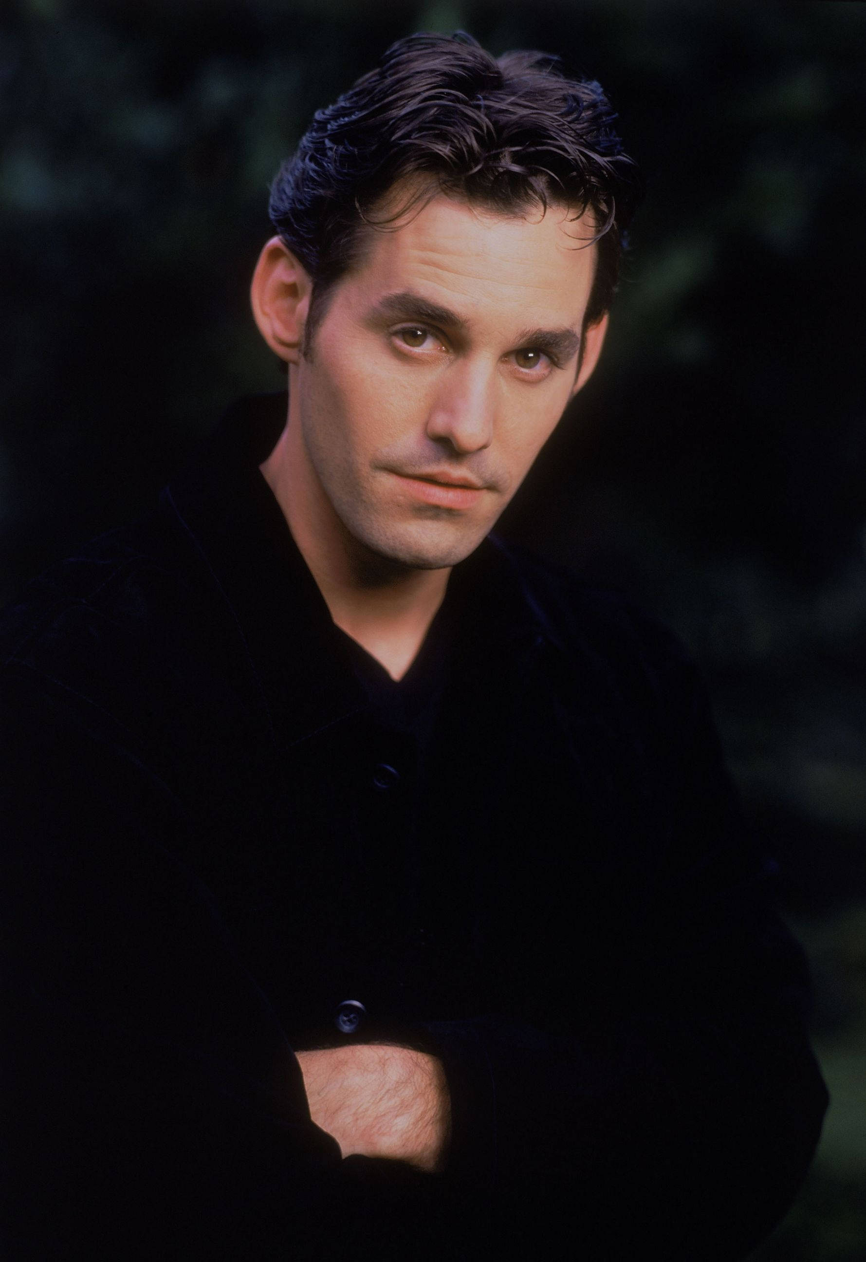 Nicholas Brendon 桌布 壁紙 — 一個穿著黑色外套的男人雙臂交叉站立，背景黑暗模糊，直視鏡頭，表情嚴肅。
