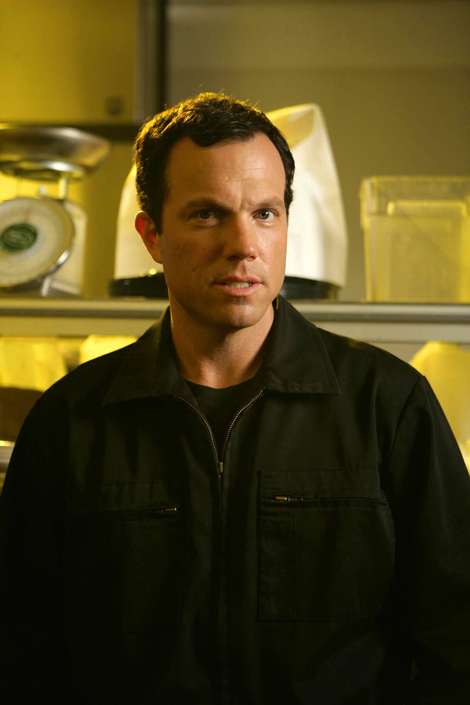 adam baldwin 桌布 壁紙 — 一個穿著黑色工作外套的男人站在一個溫暖照明的廚房裡，背景有黃色櫥櫃和一個秤。