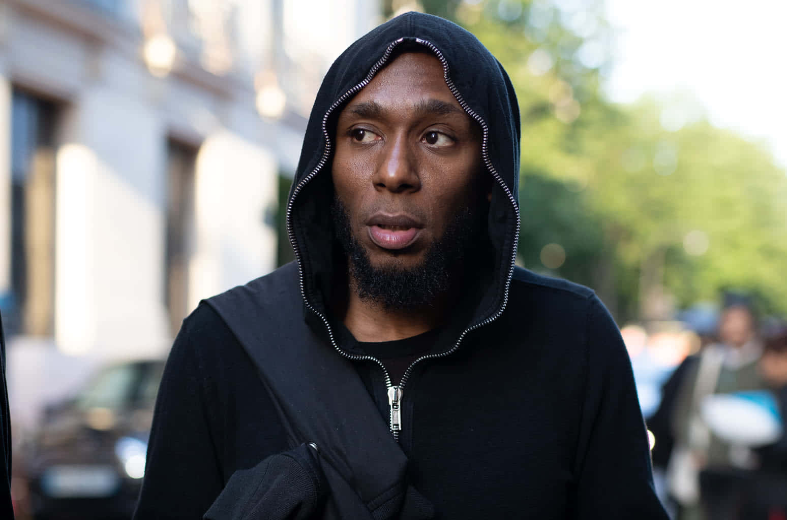Yasiin Bey 桌布 壁紙 — 一個穿著黑色拉鍊連帽衫（帶銀色拉鍊）的男人站在陽光明媚的城市街道上，若有所思地望向一旁。