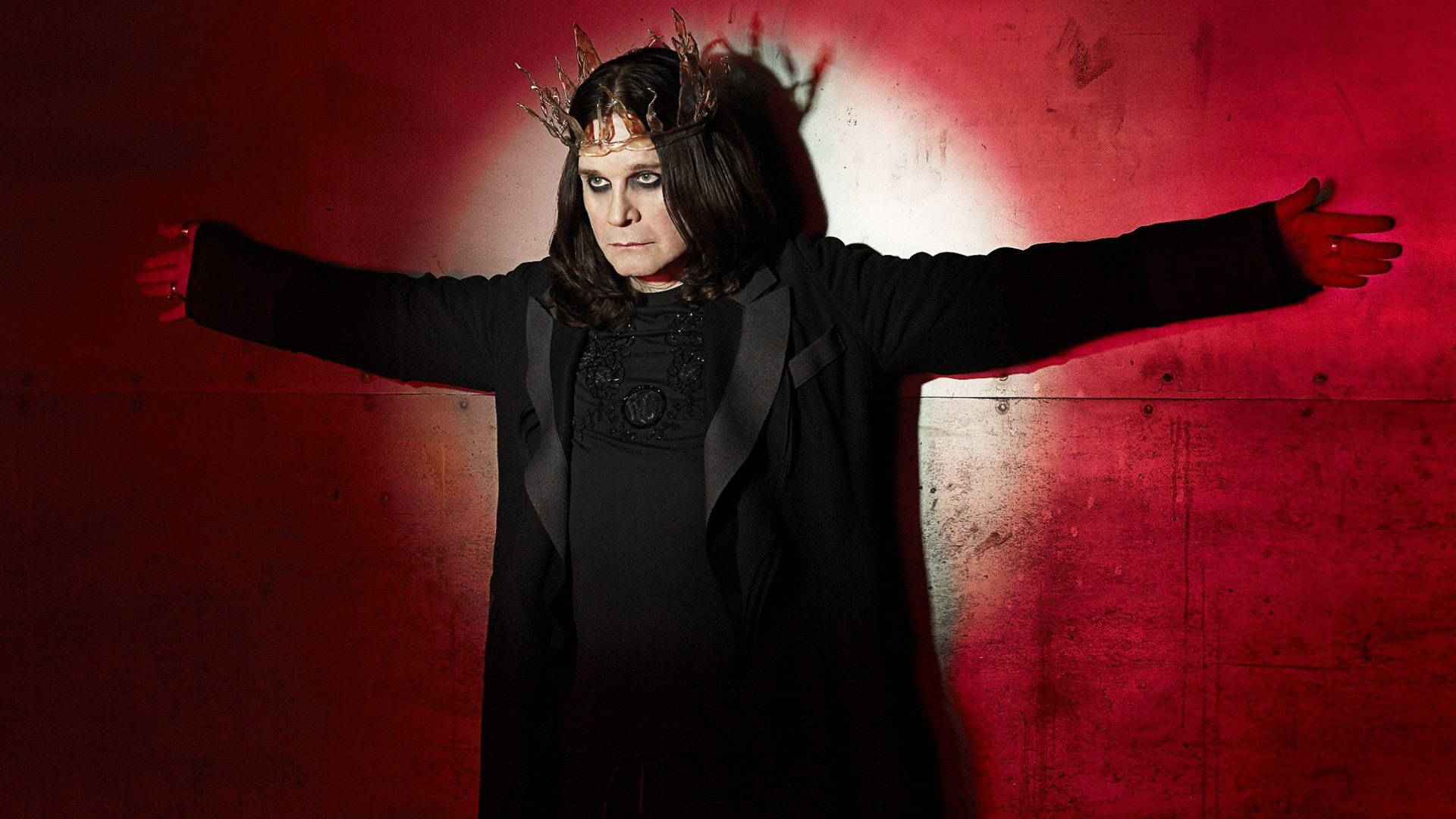 Ozzy Osbourne 桌布 壁紙 — 一個穿著黑色服裝和帶刺皇冠的人，在戲劇性的聚光燈下，雙臂伸展，面向鮮紅色的紋理牆壁。