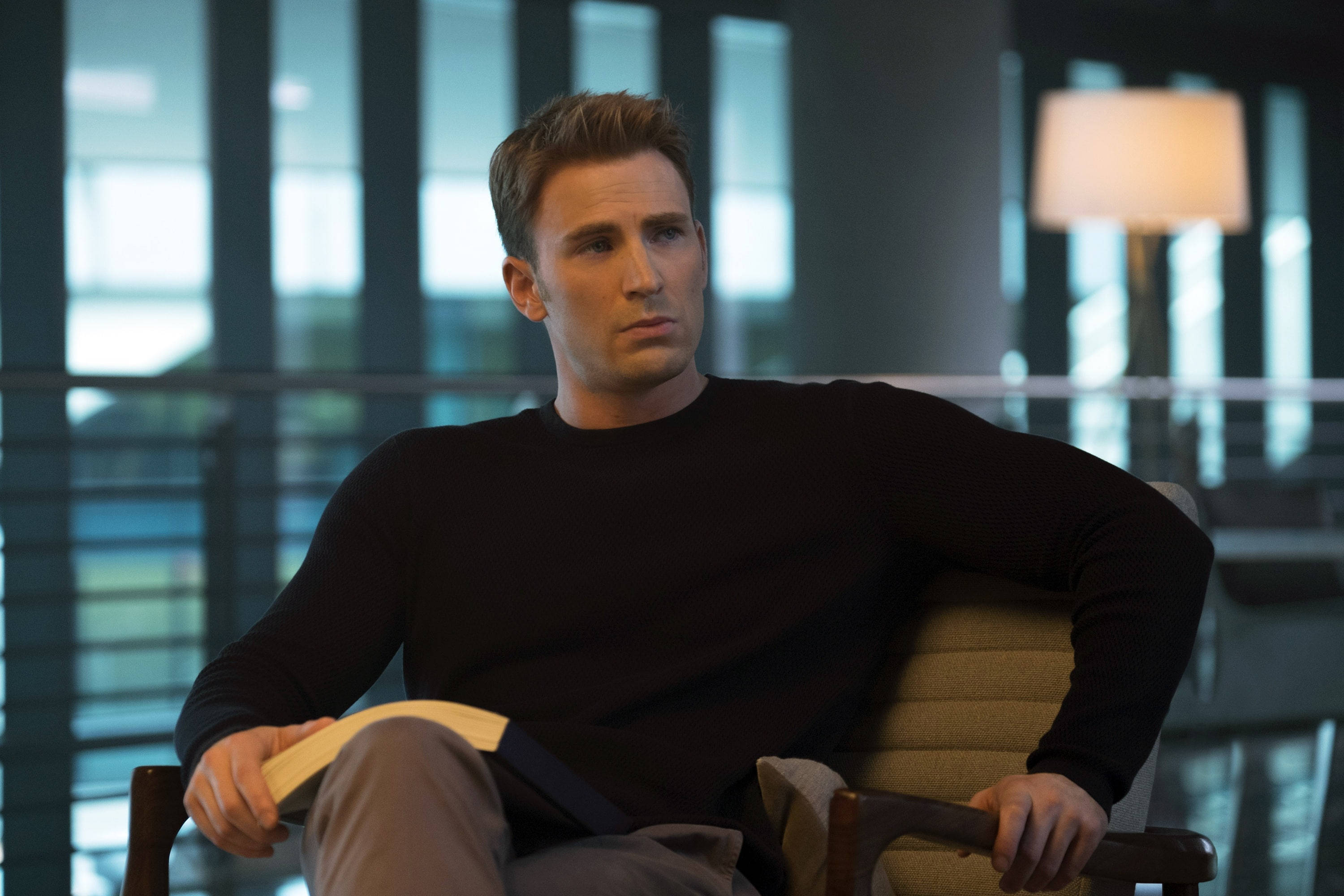 Marvel Captain America 桌布 壁紙 — 一個穿著黑色毛衣的男人坐在椅子上，拿著一本書，在柔和燈光的現代室內環境中沉思。
