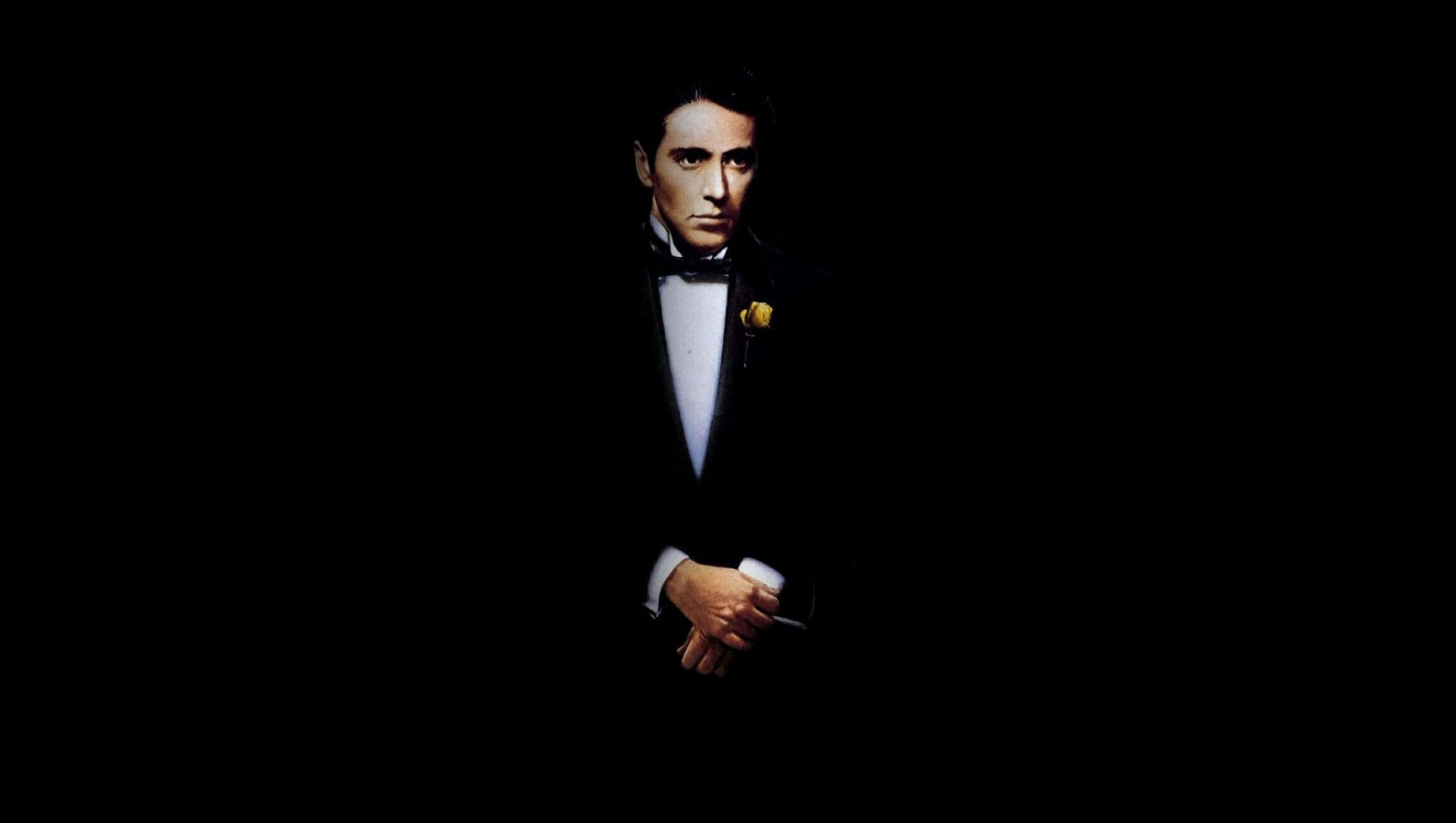 Michael Corleone 桌布 壁紙 — 一個穿著黑色燕尾服、佩戴黃色襟花的男人，在漆黑的背景下莊嚴地站立。