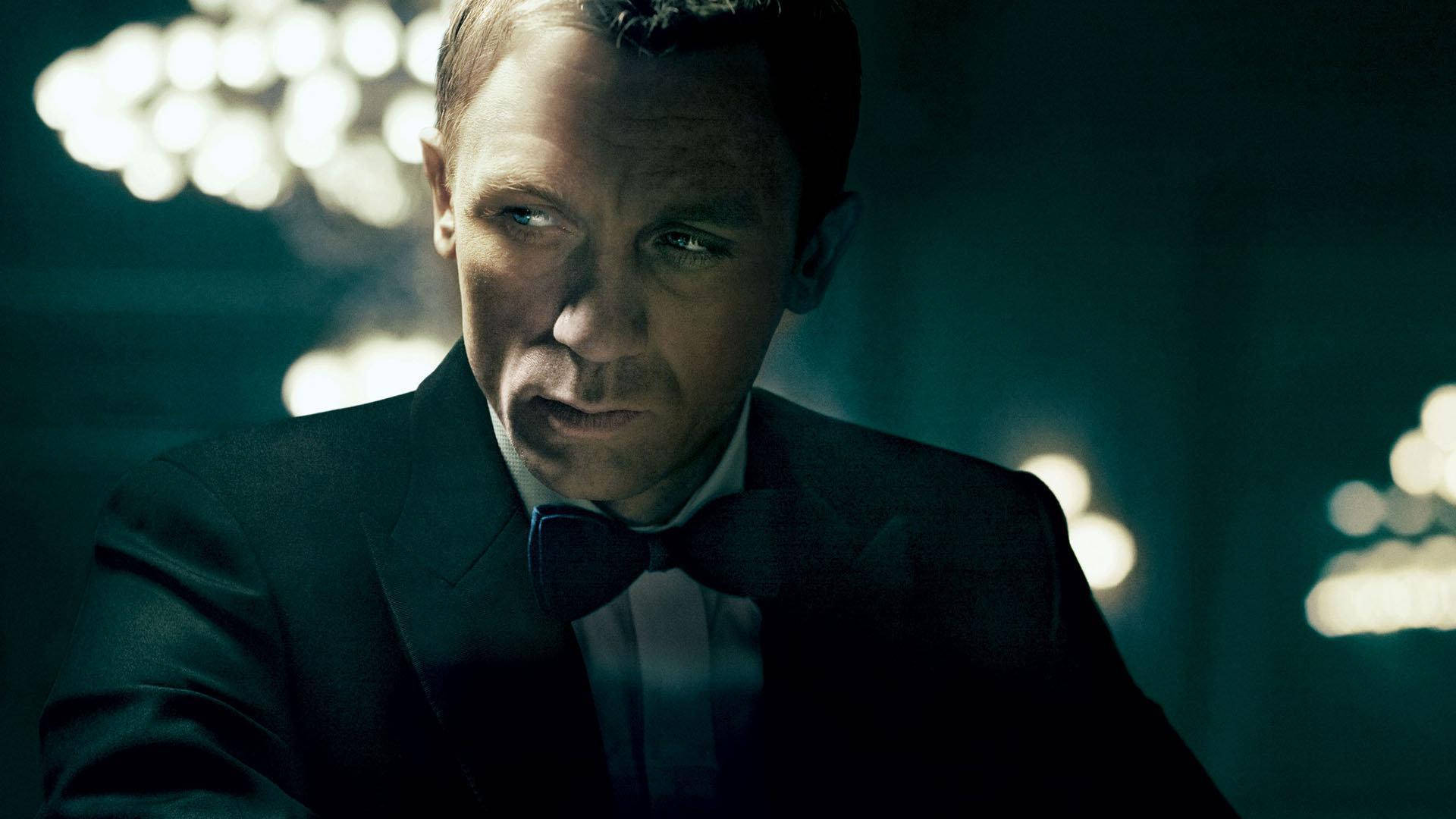 Daniel Craig 桌布 壁紙 — 一個穿著黑色燕尾服和領結的男人在昏暗的房間裡凝視著，身後有發光的水晶吊燈。