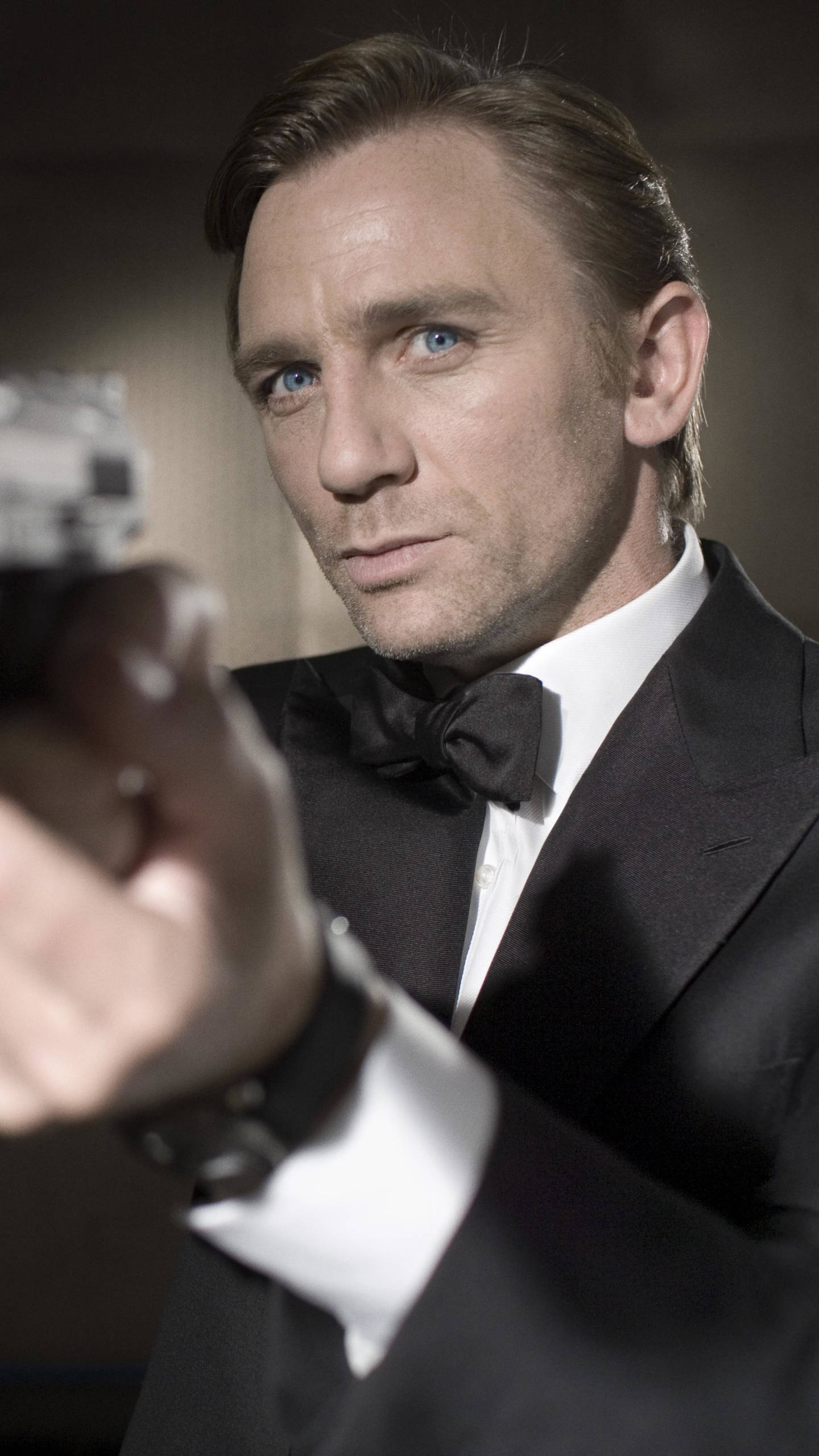 Daniel Craig 桌布 壁紙 — 一個穿著黑色燕尾服的男人在昏暗的房間裡用強烈的藍眼睛瞄準槍。
