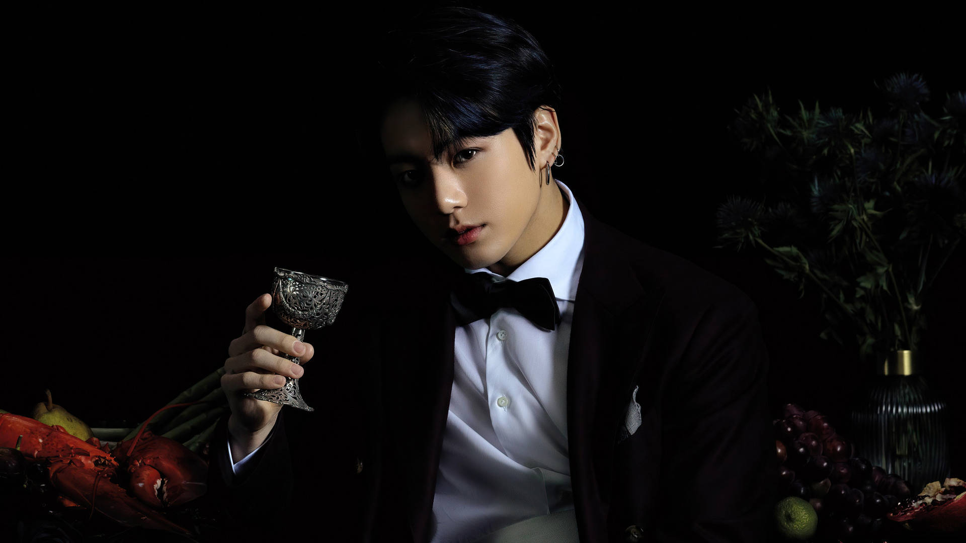 Jungkook 桌布 壁紙 — 一個穿著黑色燕尾服的男人在黑暗背景下拿著一個華麗的玻璃杯，旁邊有水果和花朵。