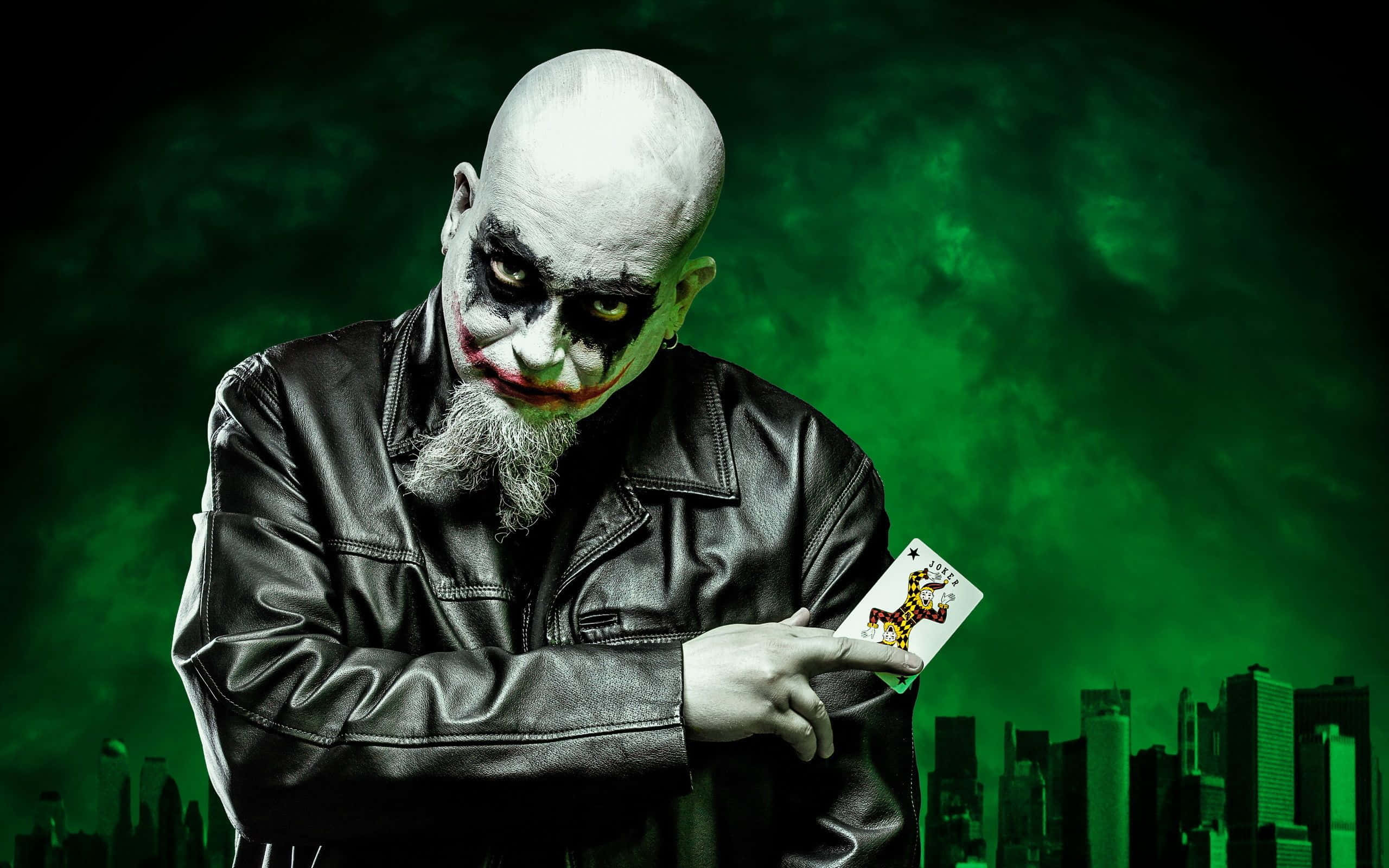Evil Joker 桌布 壁紙 — 一個穿著黑色皮夾克的威脅性小丑，在發光的綠色城市景觀背景下拿著一張小丑牌。
