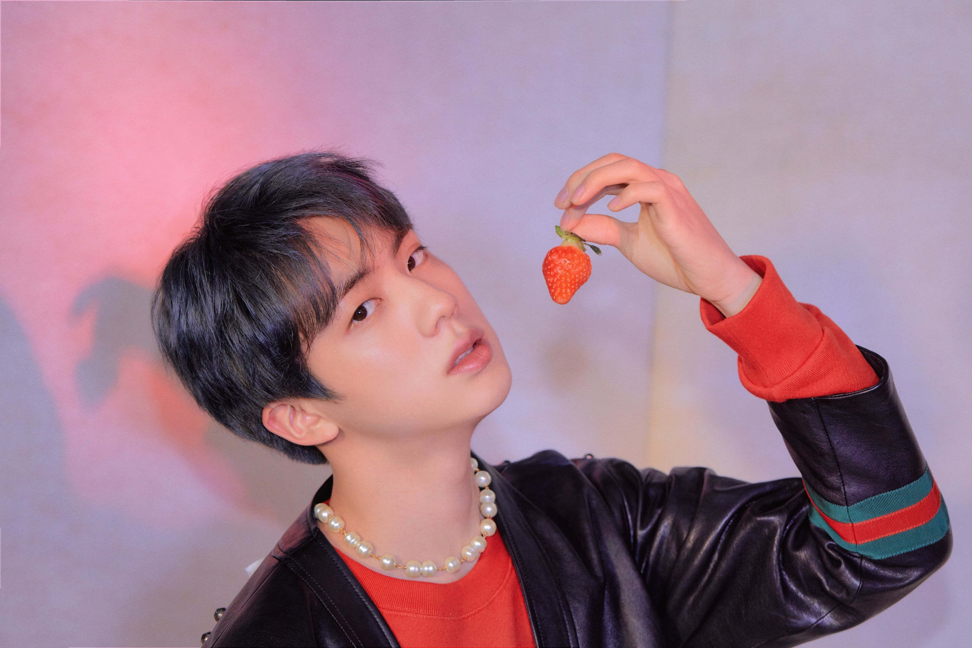 Jin 桌布 壁紙 — 一個穿著黑色皮夾克的年輕男子，在柔和的粉紅和紫色漸變背景前，將草莓靠近臉部。