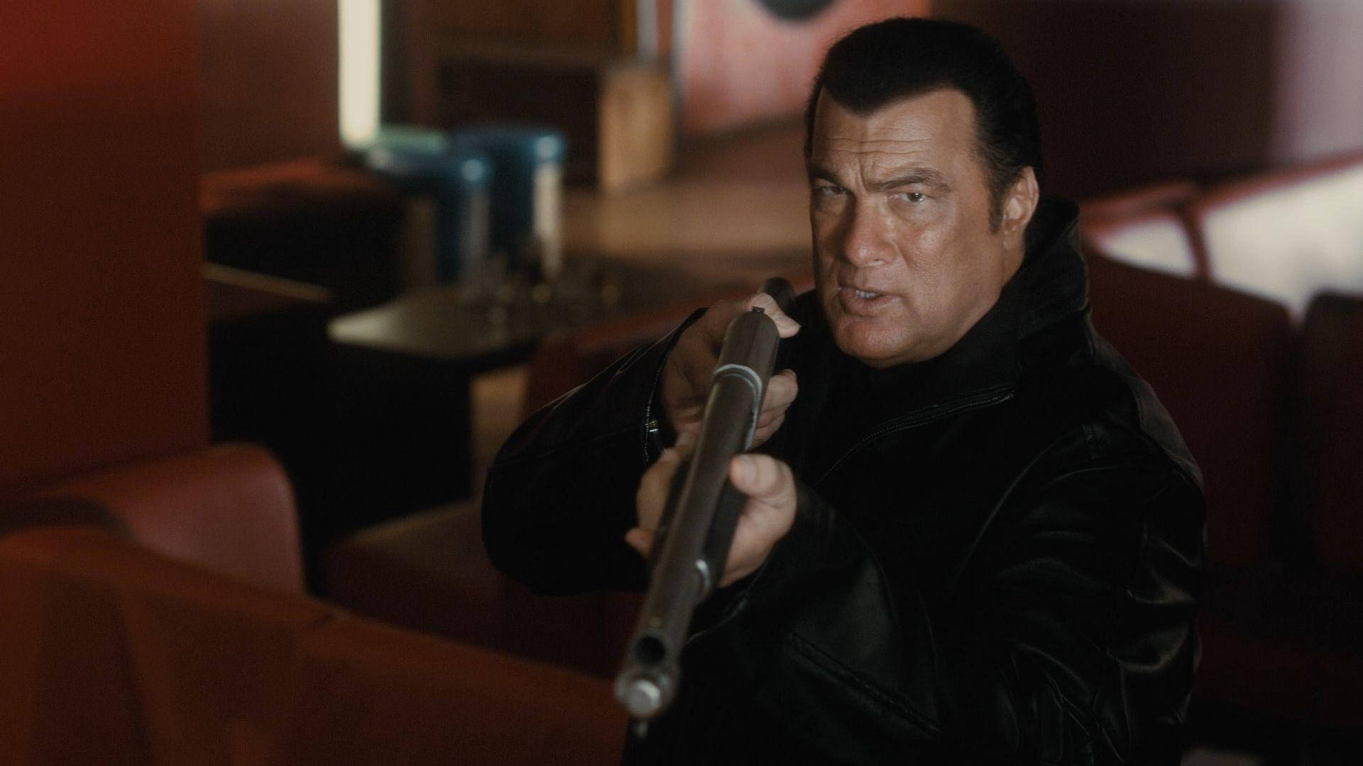Steven Seagal 桌布 壁紙 — 一個穿著黑色皮夾克的男人，在燈光昏暗、有紅色卡座的餐廳裡，全神貫注地瞄準一把散彈槍。