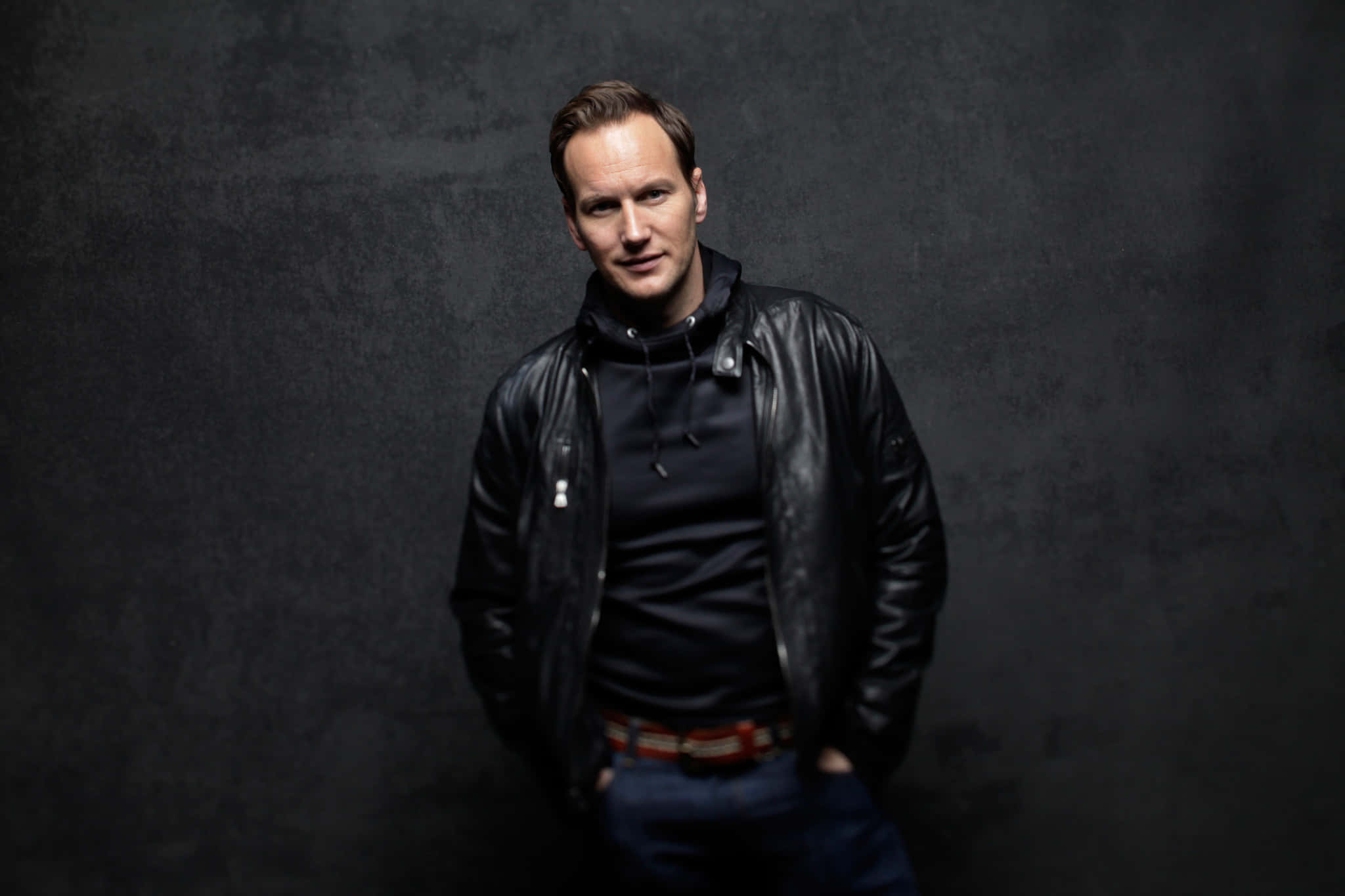 Patrick Wilson 桌布 壁紙 — 一個穿著黑色皮夾克的男人，雙手插在口袋裡，自信地站在有紋理的深灰色攝影棚背景前。