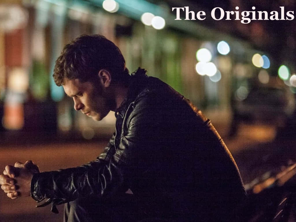Vampire Diaries 桌布 壁紙 — 一個穿著黑色皮夾克的男人晚上獨自坐在長椅上，被柔和的城市燈光照亮，背景是模糊的城市景觀。