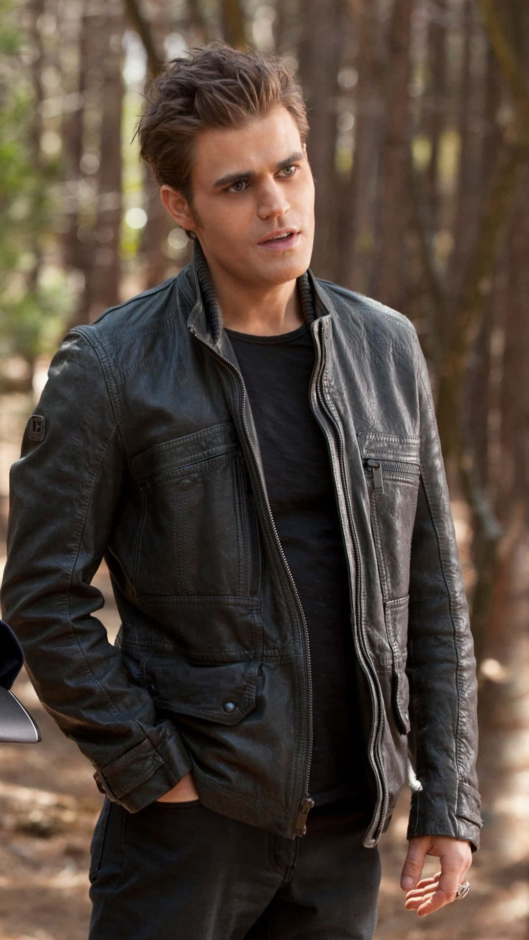 Vampire Diaries Desktop 桌布 壁紙 — 一個穿著黑色皮夾克的男人站在陽光明媚的森林中，背景的樹木模糊不清。