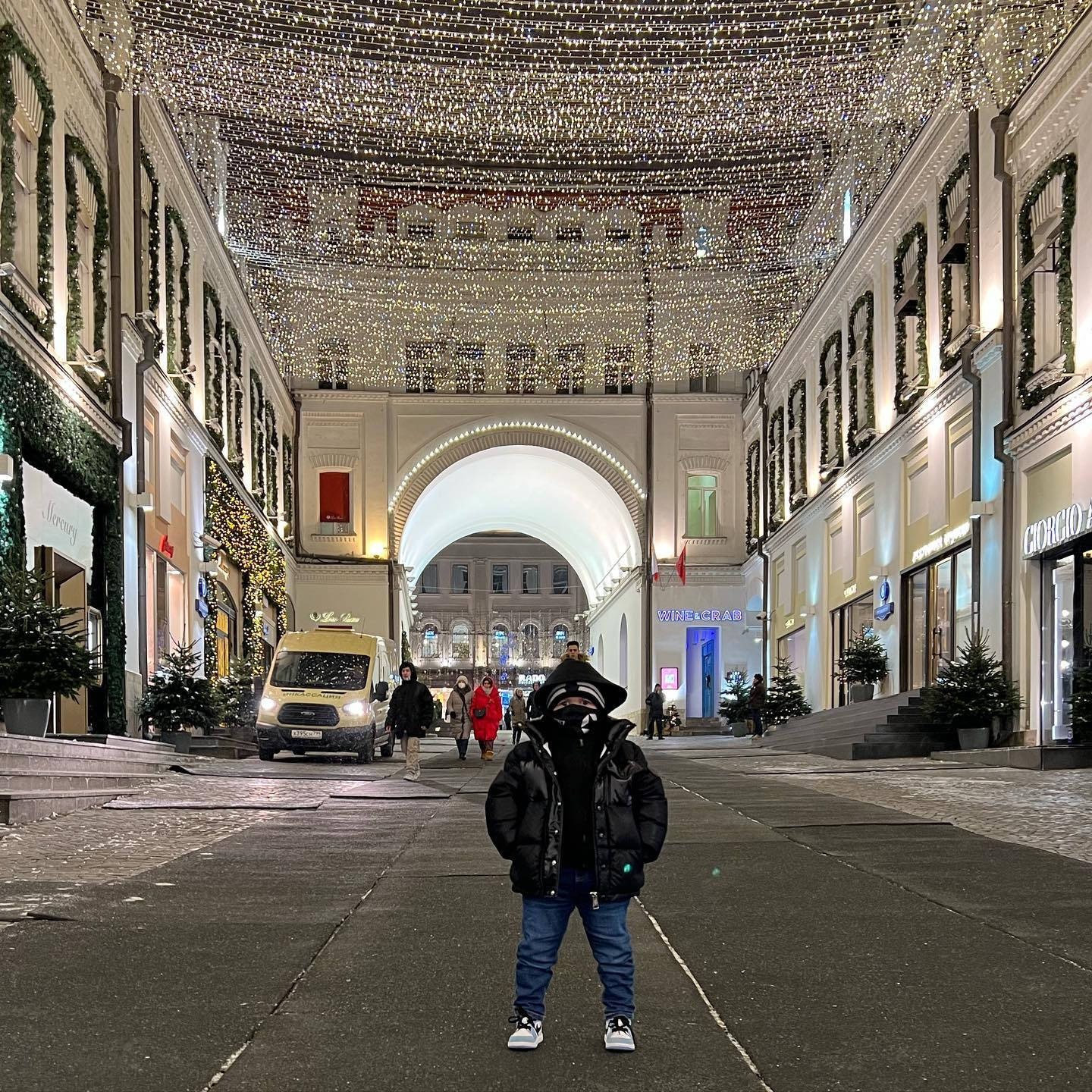 Building 桌布 壁紙 — 一個穿著黑色羽絨外套的孩子，在夜晚宏偉的城市拱廊下，站在耀眼的聖誕燈飾天篷中央。
