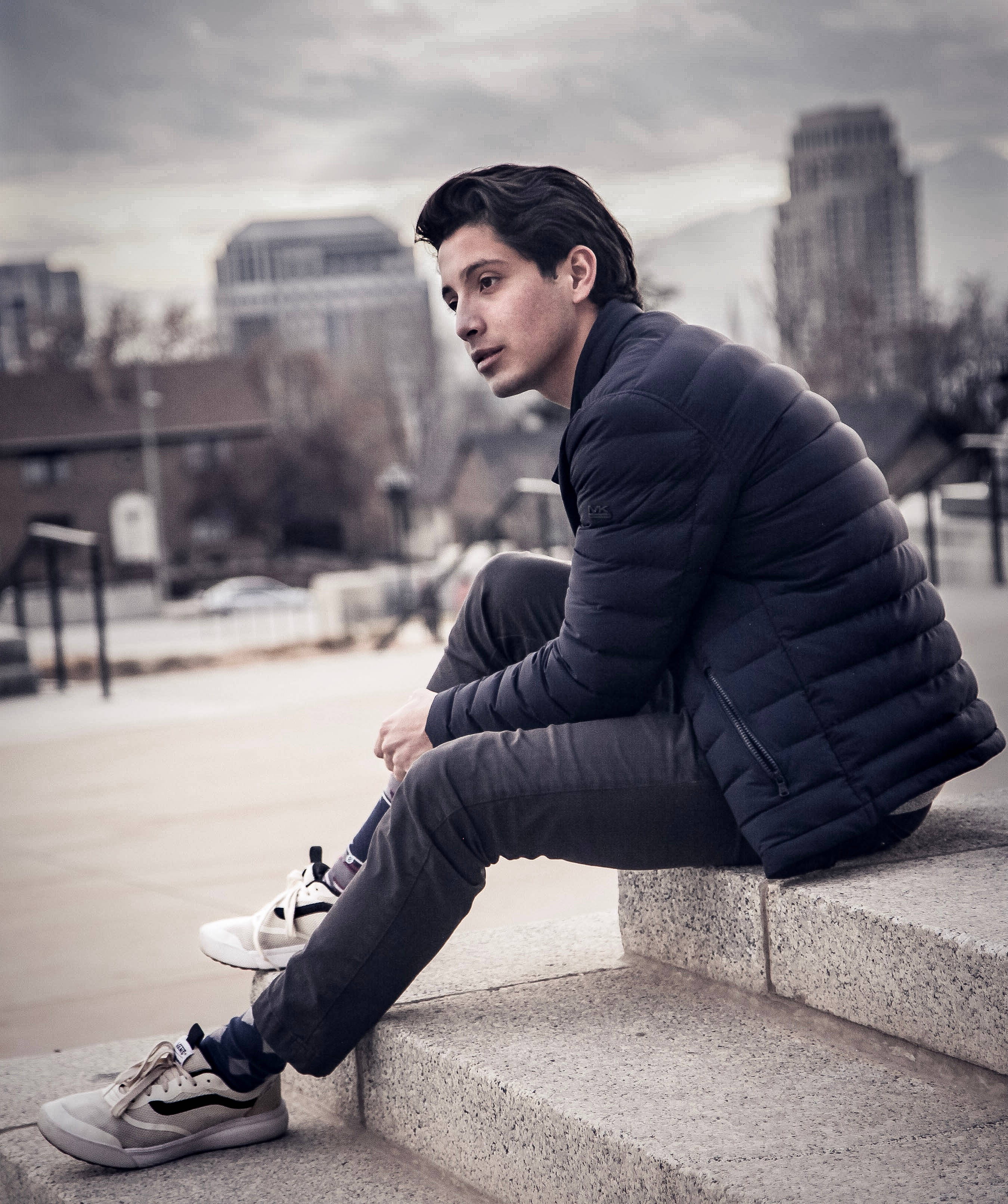 cool shoe 桌布 壁紙 — 一個穿著黑色羽絨外套的年輕男子，在陰天之下，沉思地坐在石階上，背景是柔和的城市天際線。
