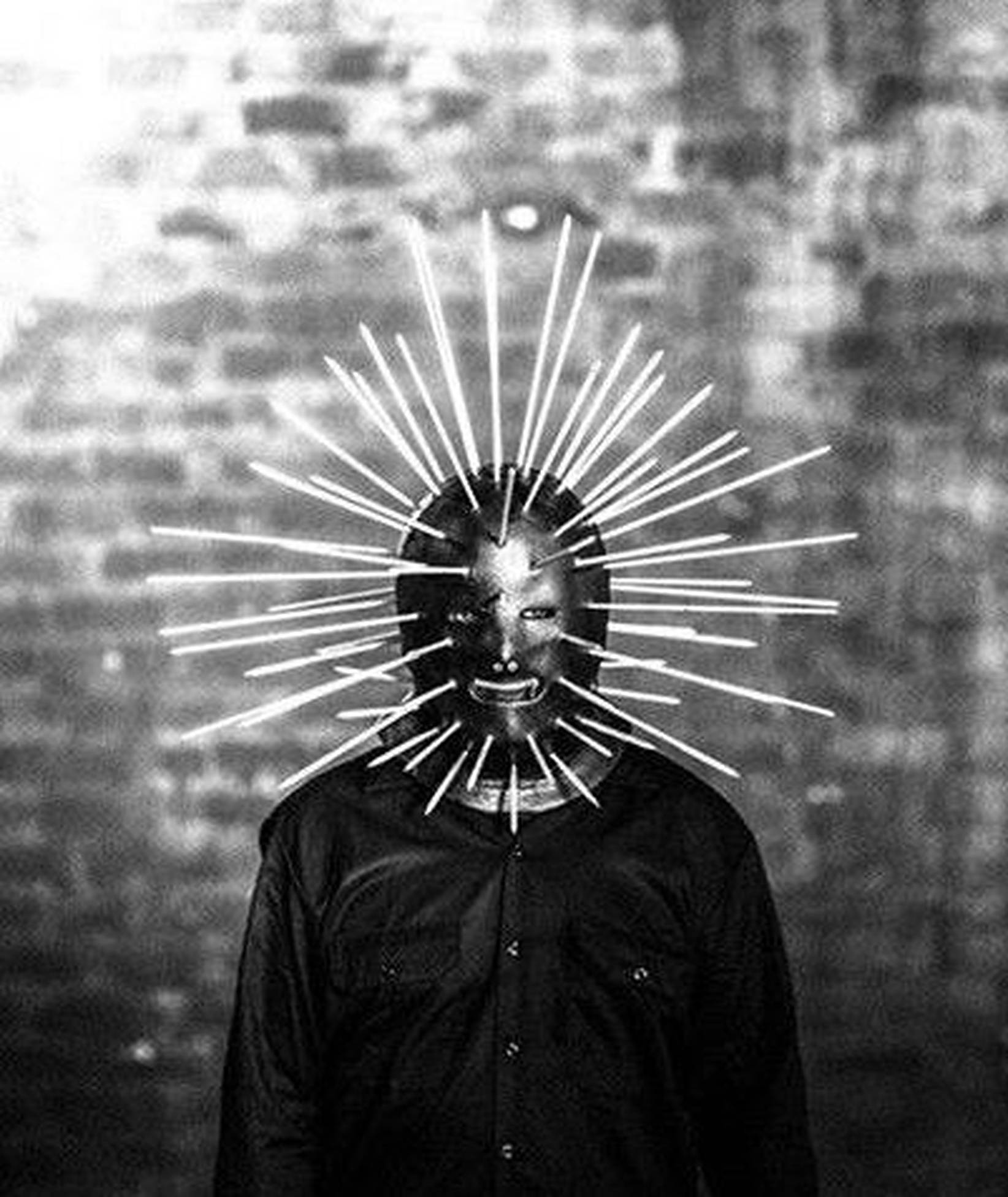 craig jones 桌布 壁紙 — 一個穿著黑色襯衫的人戴著一個金屬面具，面具上有尖銳的白色尖刺向外輻射，背景是磚牆。