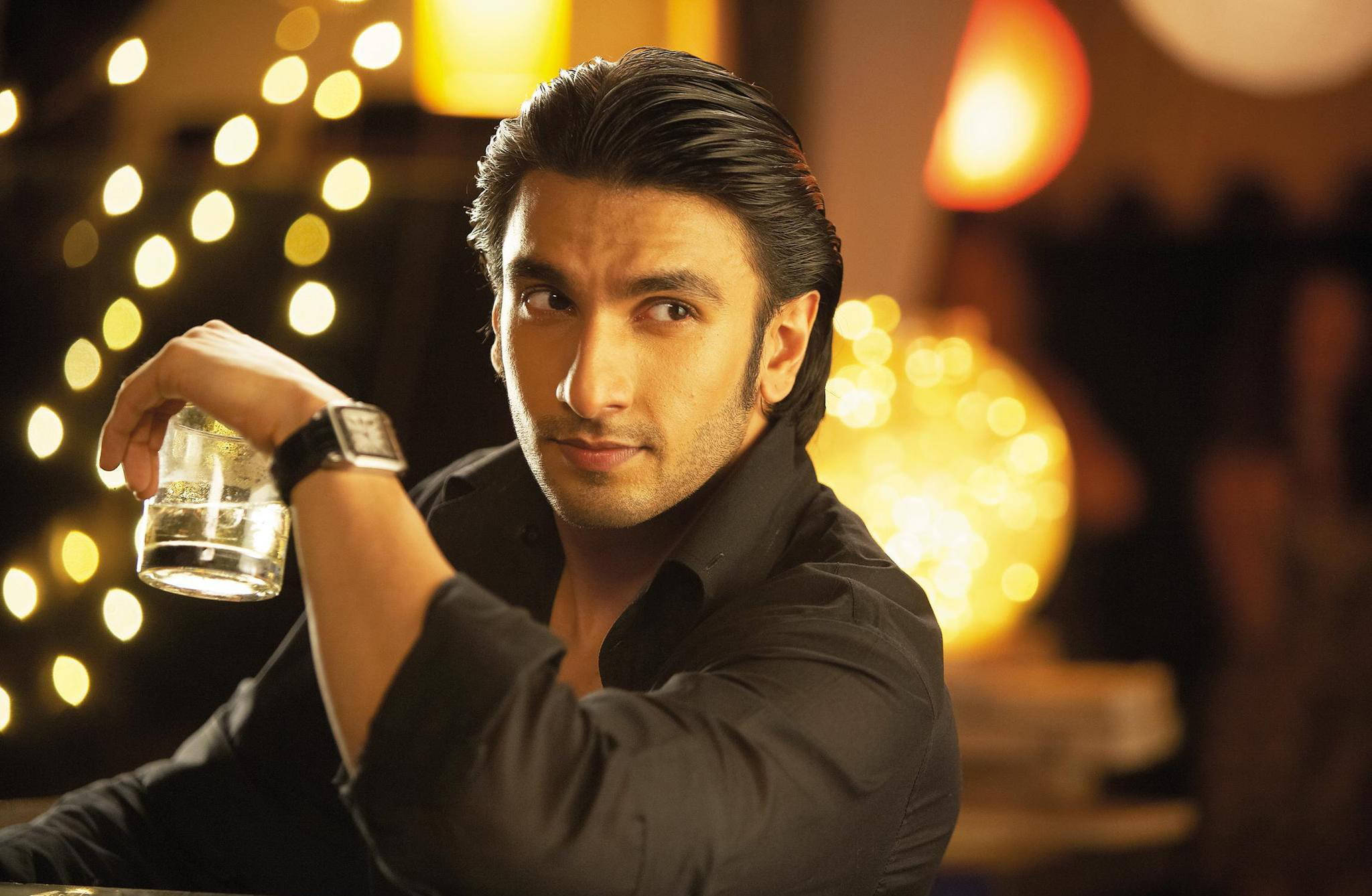 Ranveer Singh 桌布 壁紙 — 一個穿著黑色襯衫的男人在一個溫暖照明的酒吧裡，背景有串燈和發光球體，手持一杯水。