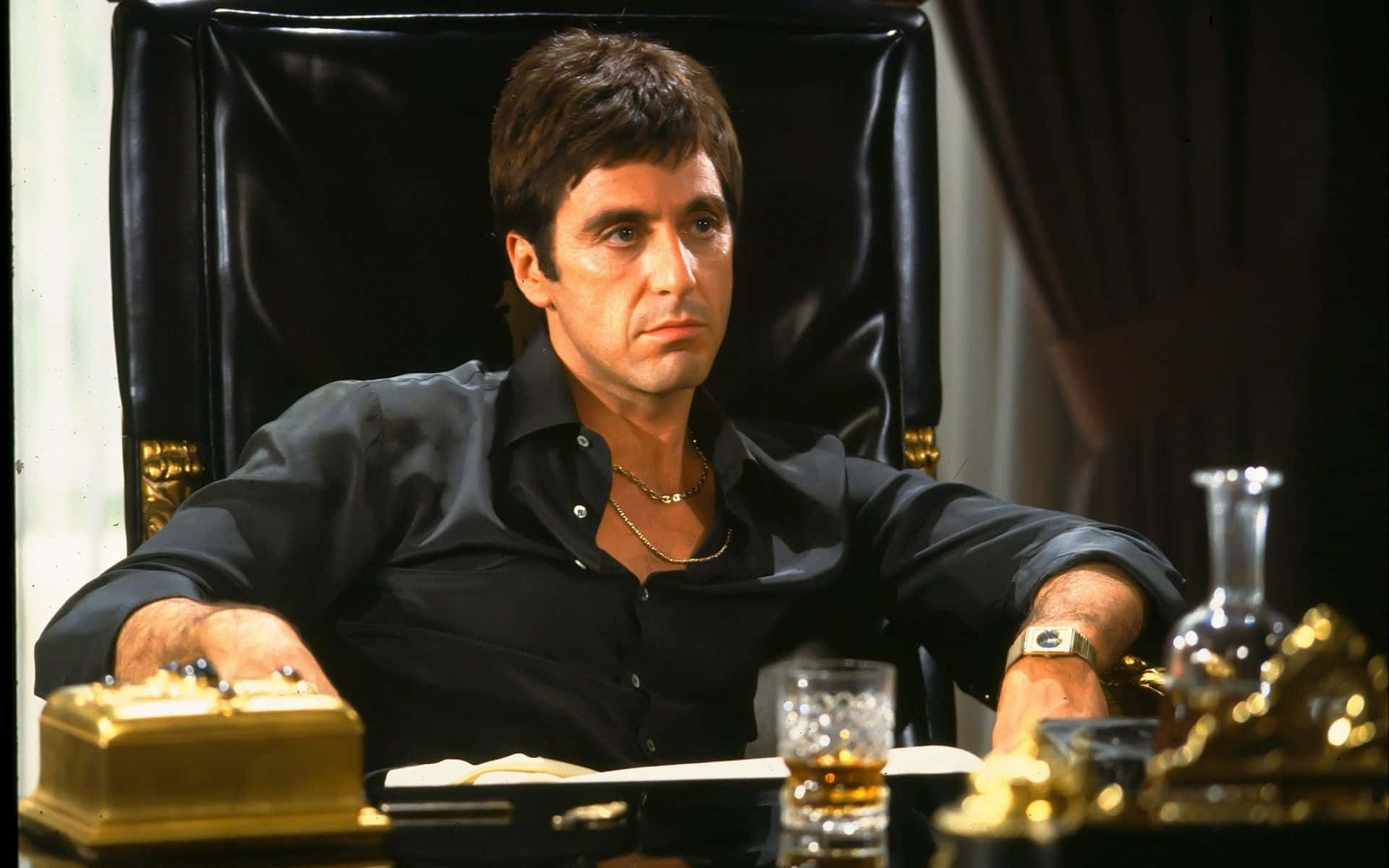 Scarface Desktop 桌布 壁紙 — 一個穿著黑色襯衫的男人坐在一張帶有金色裝飾的書桌後的皮椅上，桌上有一杯威士忌。