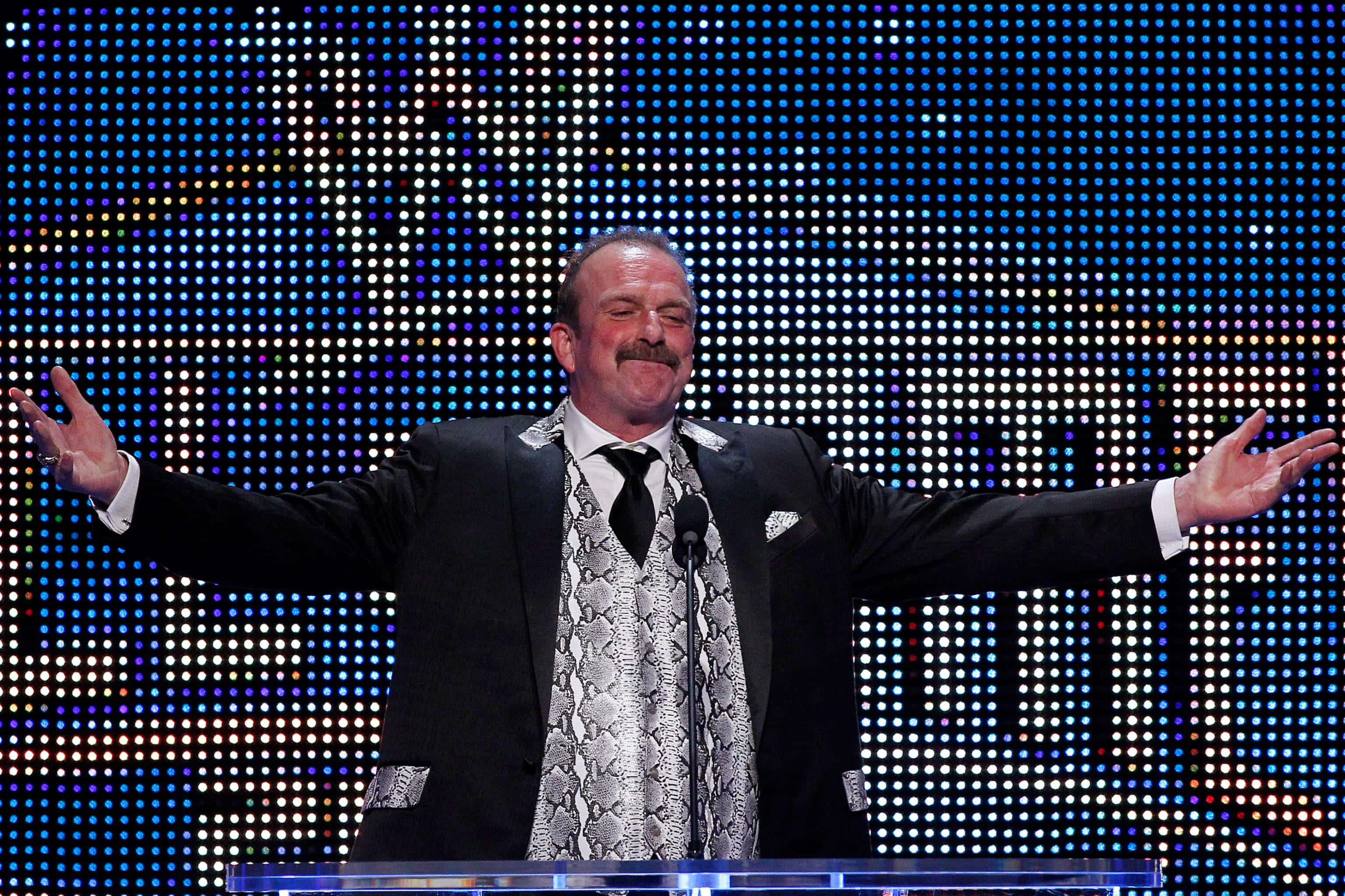 WWE 桌布 壁紙 — 一個穿著黑色西裝、打著蛇皮領帶的男人站在講台上，雙臂張開，背景是發光的藍色LED點陣牆。