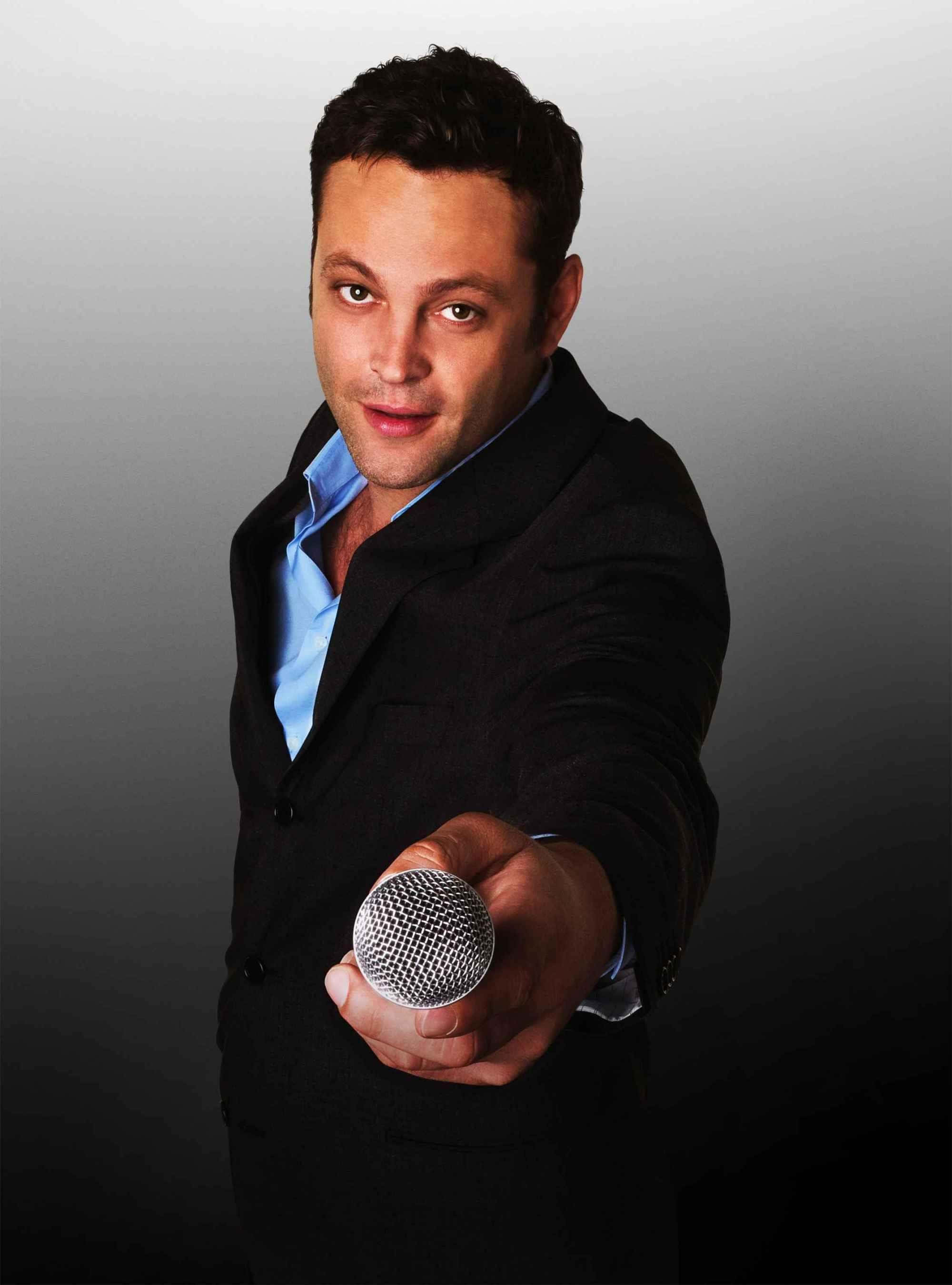 Vince Vaughn 桌布 壁紙 — 一個穿著黑色西裝外套和藍色襯衫的男人，在灰色漸層背景前，將麥克風朝向觀眾。