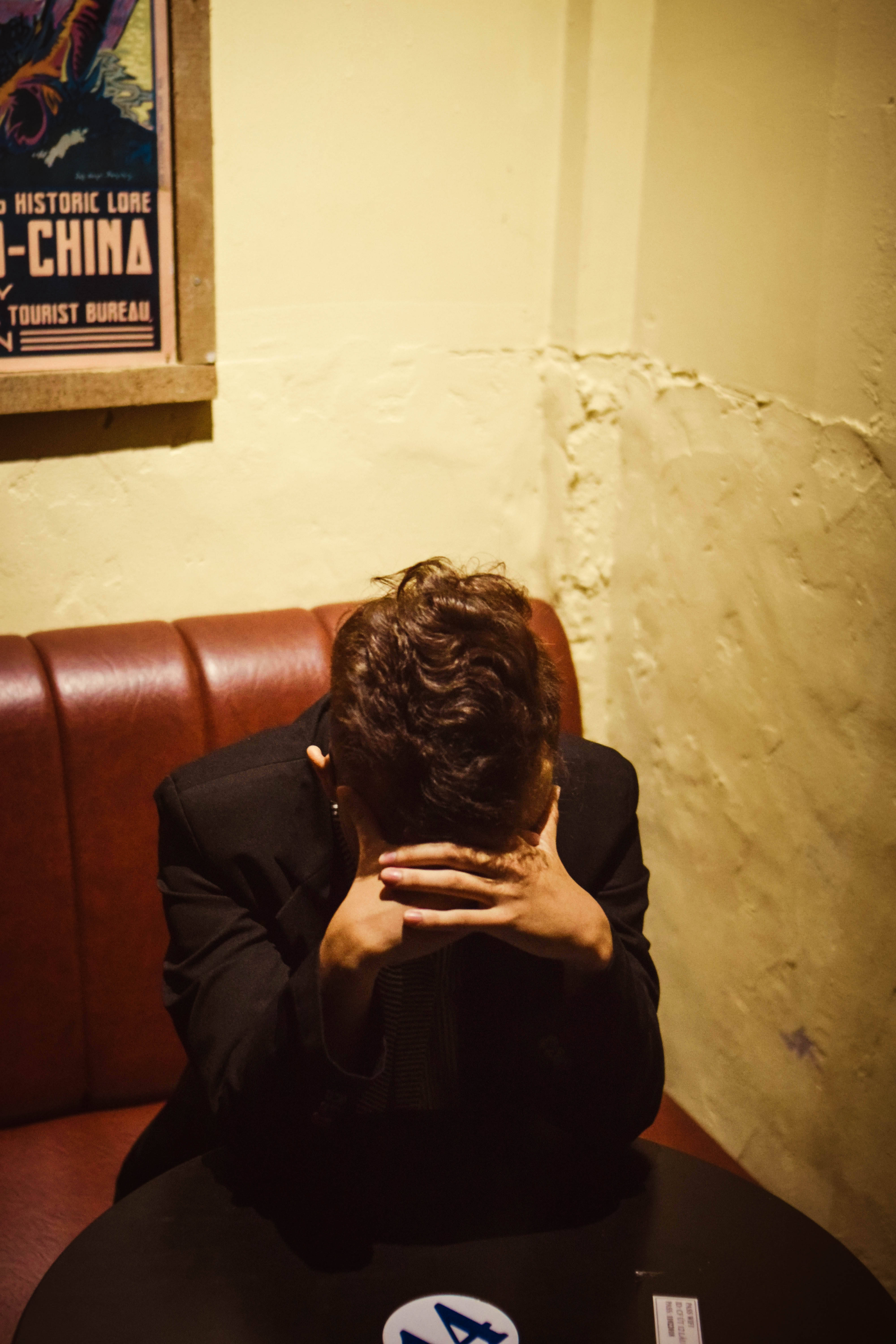 Hiding 桌布 壁紙 — 一個穿著黑色西裝的人蜷縮在紅色皮革卡座上，雙手掩面，身處燈光昏暗、溫暖的復古餐廳。