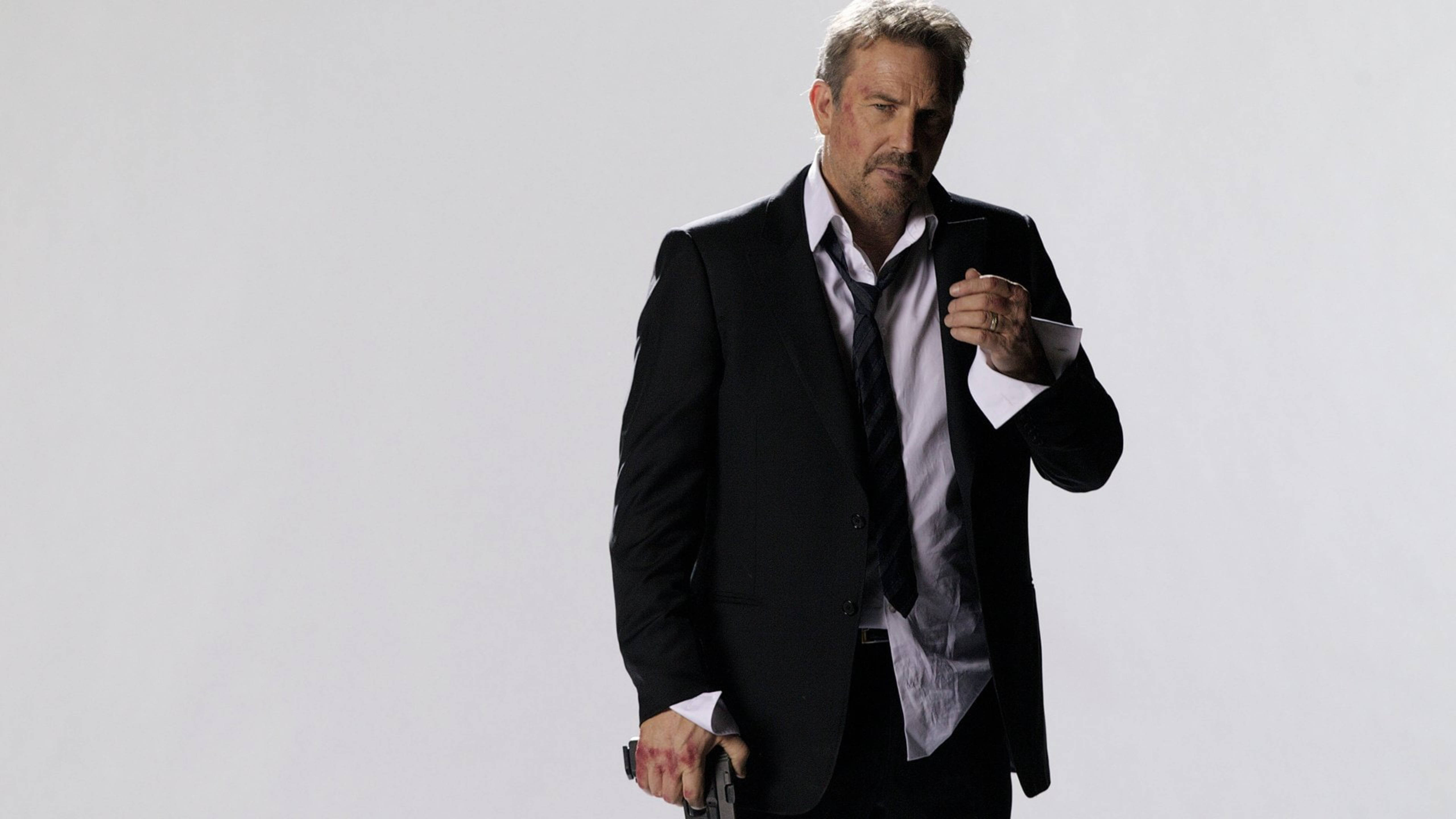 Kevin Costner 桌布 壁紙 — 一個穿著黑色西裝的男人，在純白色背景前，表情嚴肅地拿著一把槍。
