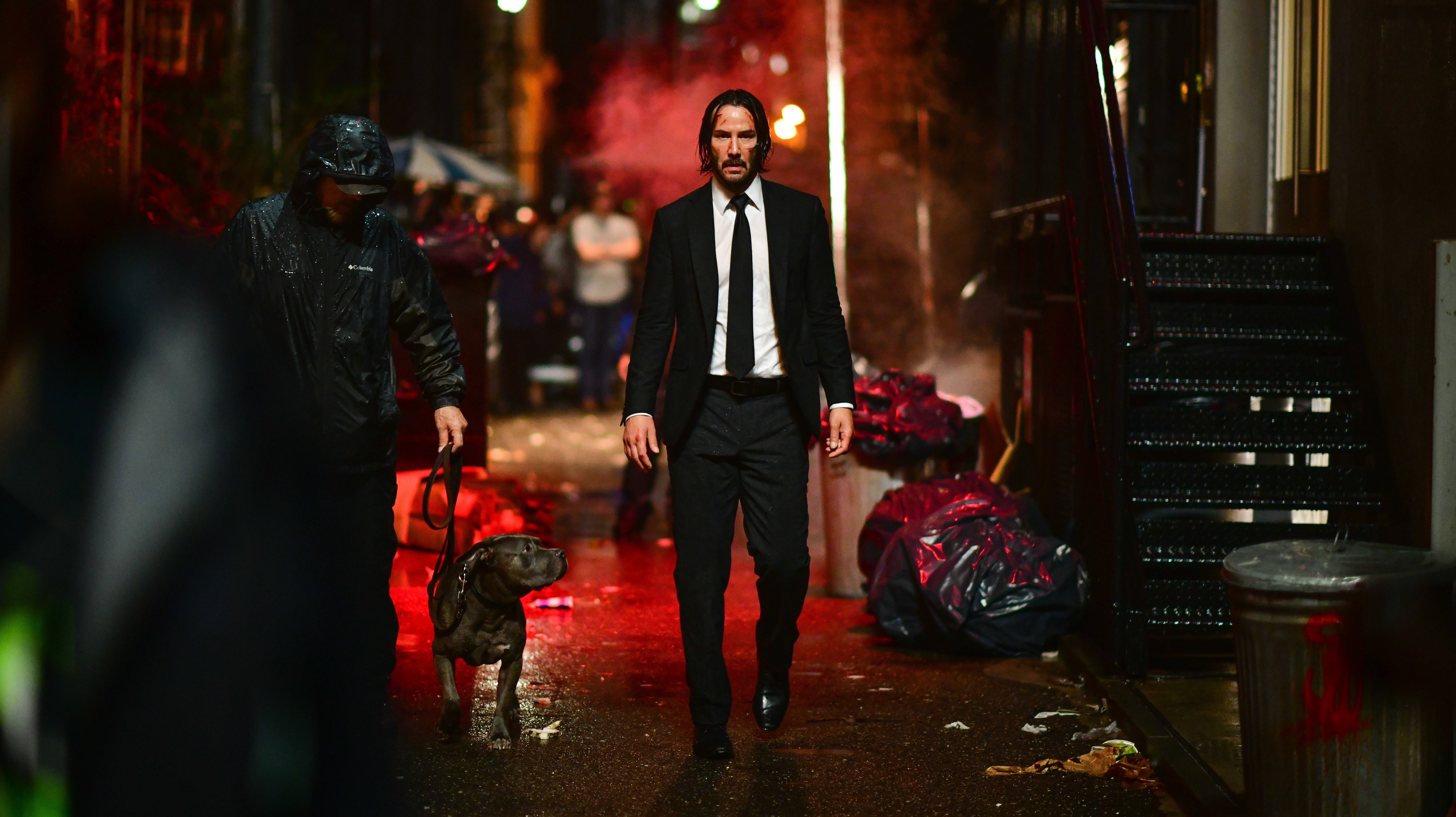 John Wick 桌布 壁紙 — 一個穿著黑色西裝的男人在夜晚沿著濕滑的巷子走，帶著一隻比特犬和一個戴帽子的同伴，在紅色霓虹燈下。