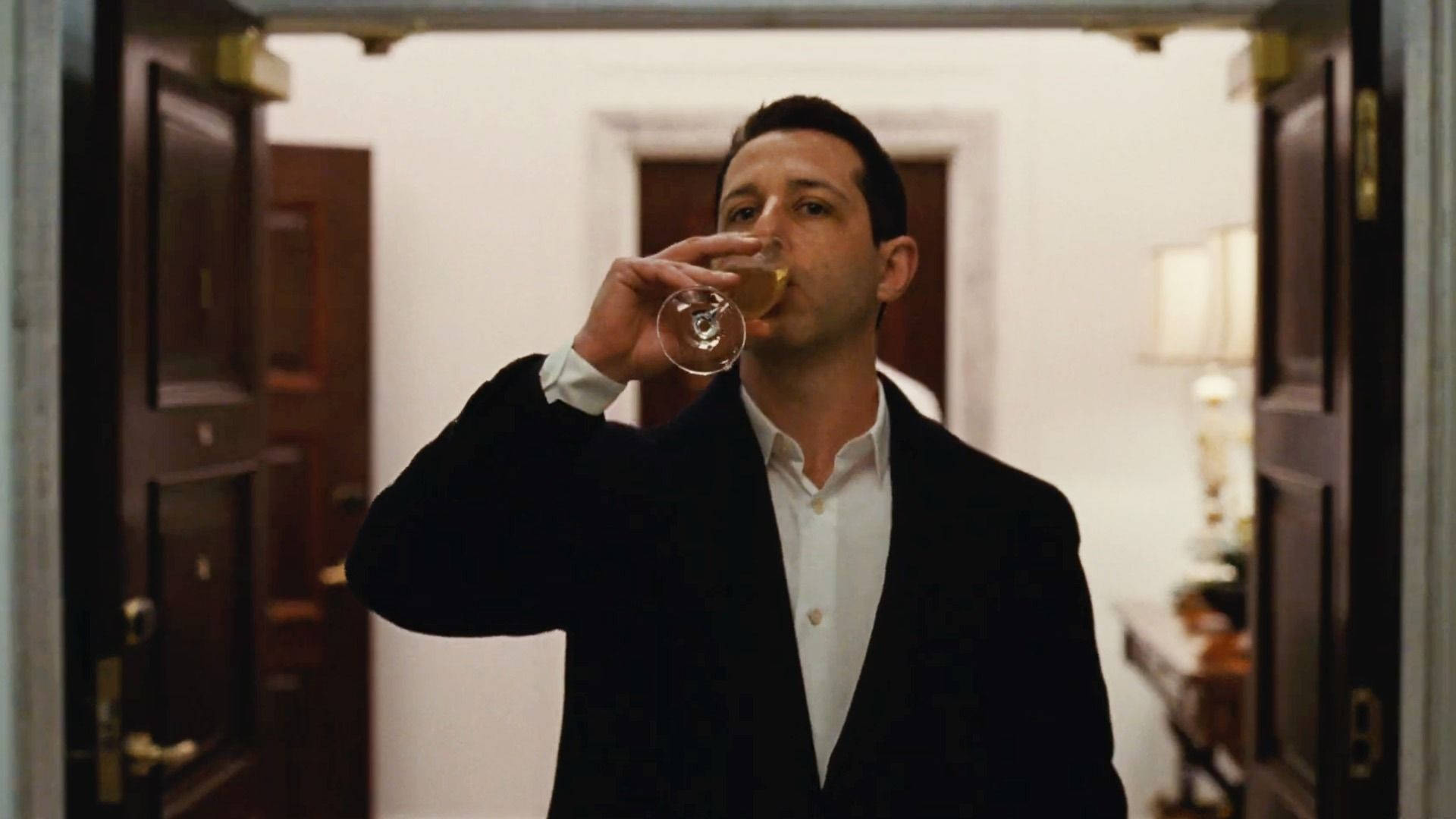Succession 桌布 壁紙 — 一個穿著黑色西裝的男人站在一個由打開的木門框著的溫暖照明走廊裡，從酒杯中啜飲。