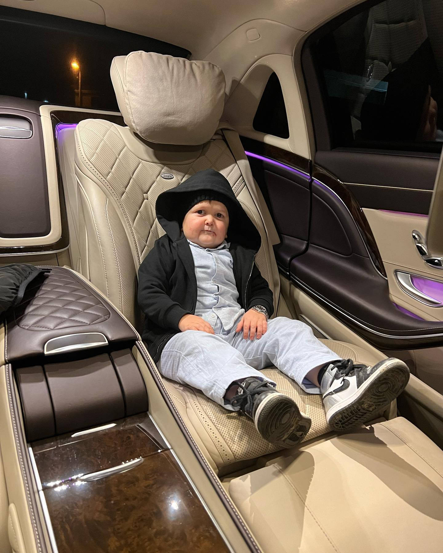 Celebrity 桌布 壁紙 — 一個穿著黑色連帽外套的幼兒自信地坐在一輛高級汽車的豪華後座，車內有紫色氛圍燈。