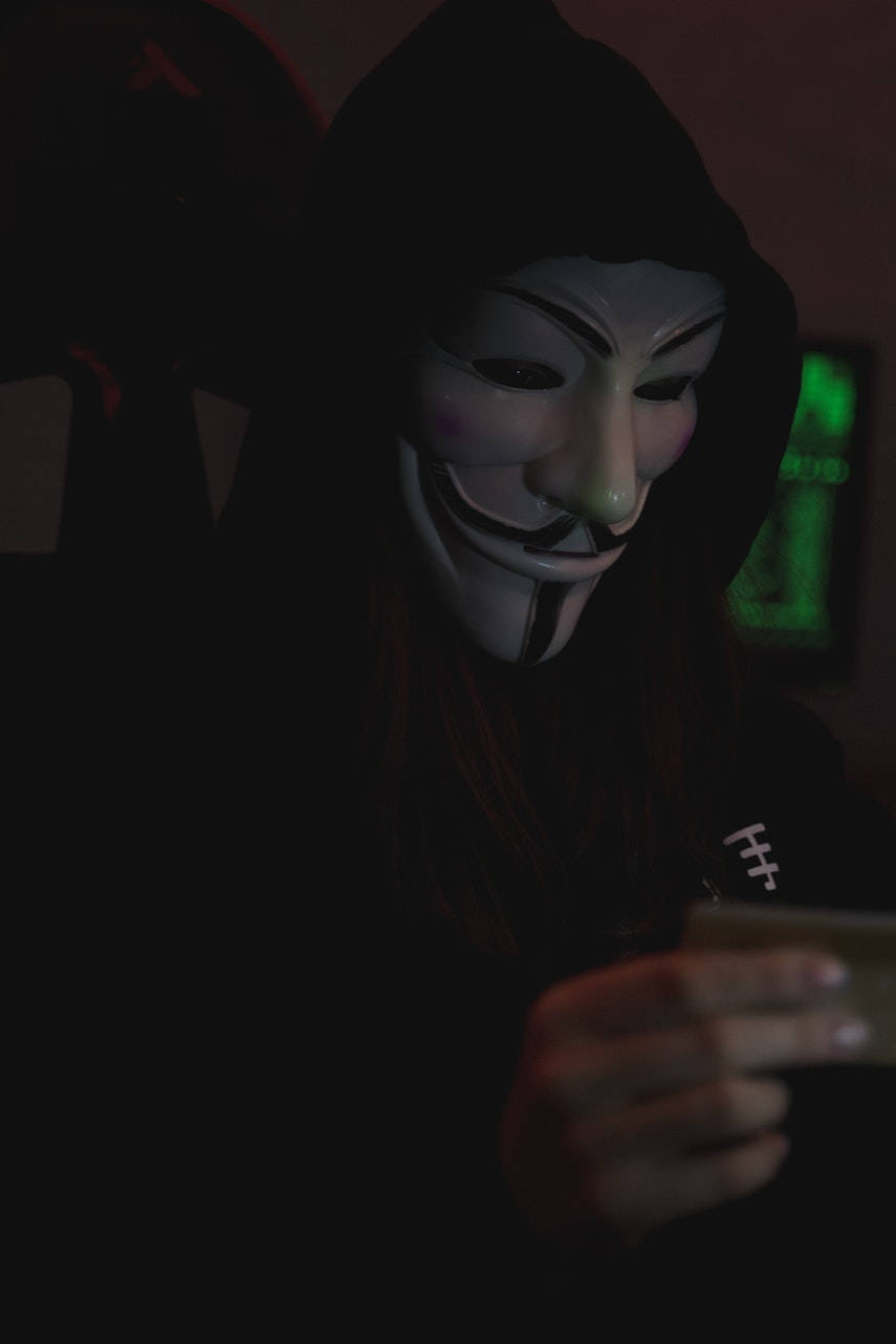hacker 3d 桌布 壁紙 — 一個穿著黑色連帽衫、戴著白色蓋伊·福克斯面具的人，在昏暗的紅光房間裡，背景有發光的綠色螢幕，手持電話。