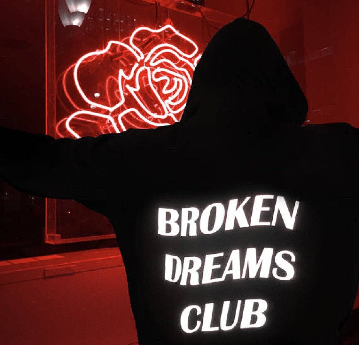 Silhouette 桌布 壁紙 — 一個穿著黑色連帽衫、背後印有「BROKEN DREAMS CLUB」字樣的人，在昏暗的房間裡張開雙臂，面向一個發光的紅色霓虹玫瑰。