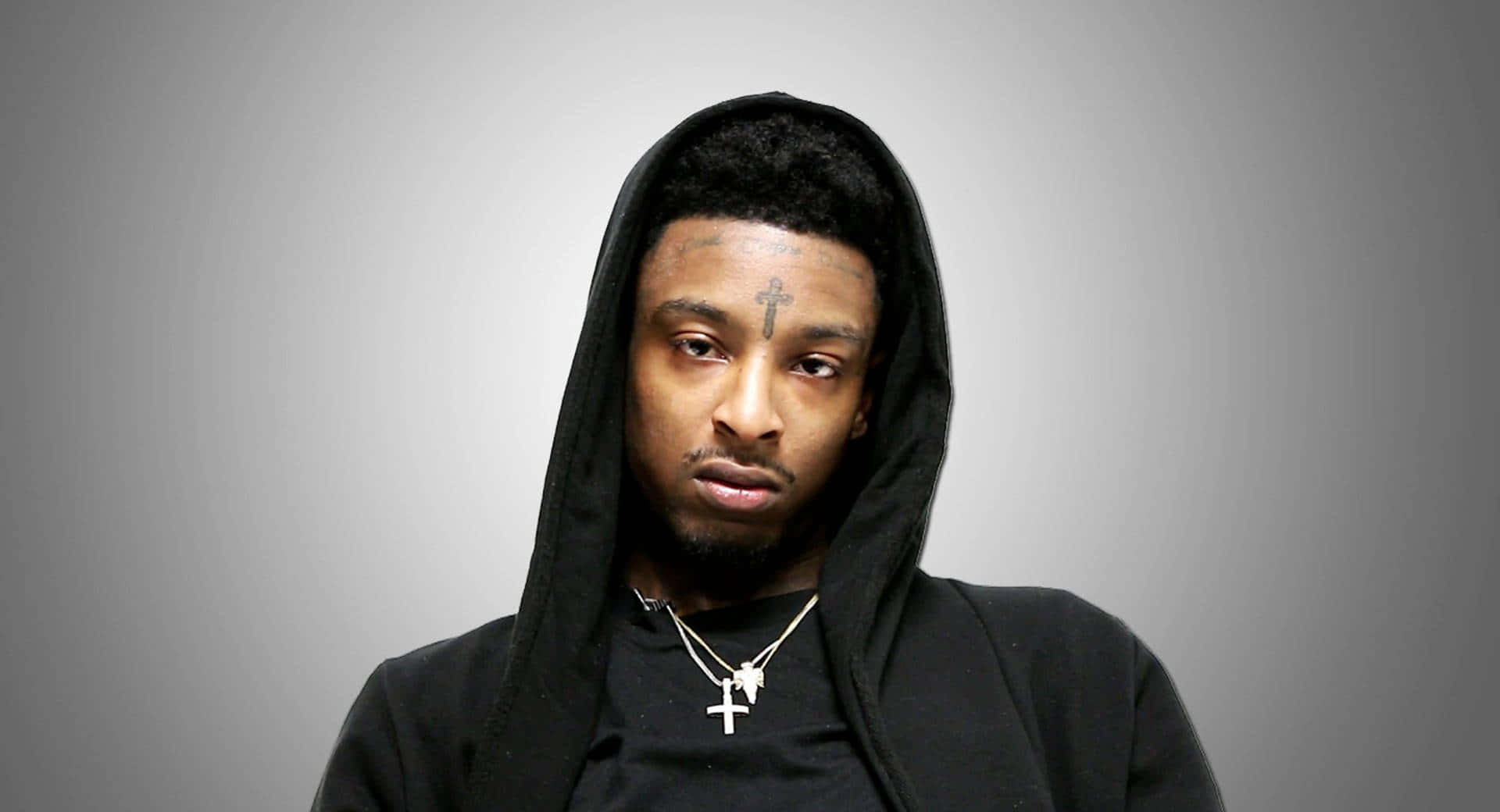 21 Savage 桌布 壁紙 — 一個穿著黑色連帽衫、額頭上有十字架刺青的男人，在純灰色背景下強烈地凝視著。