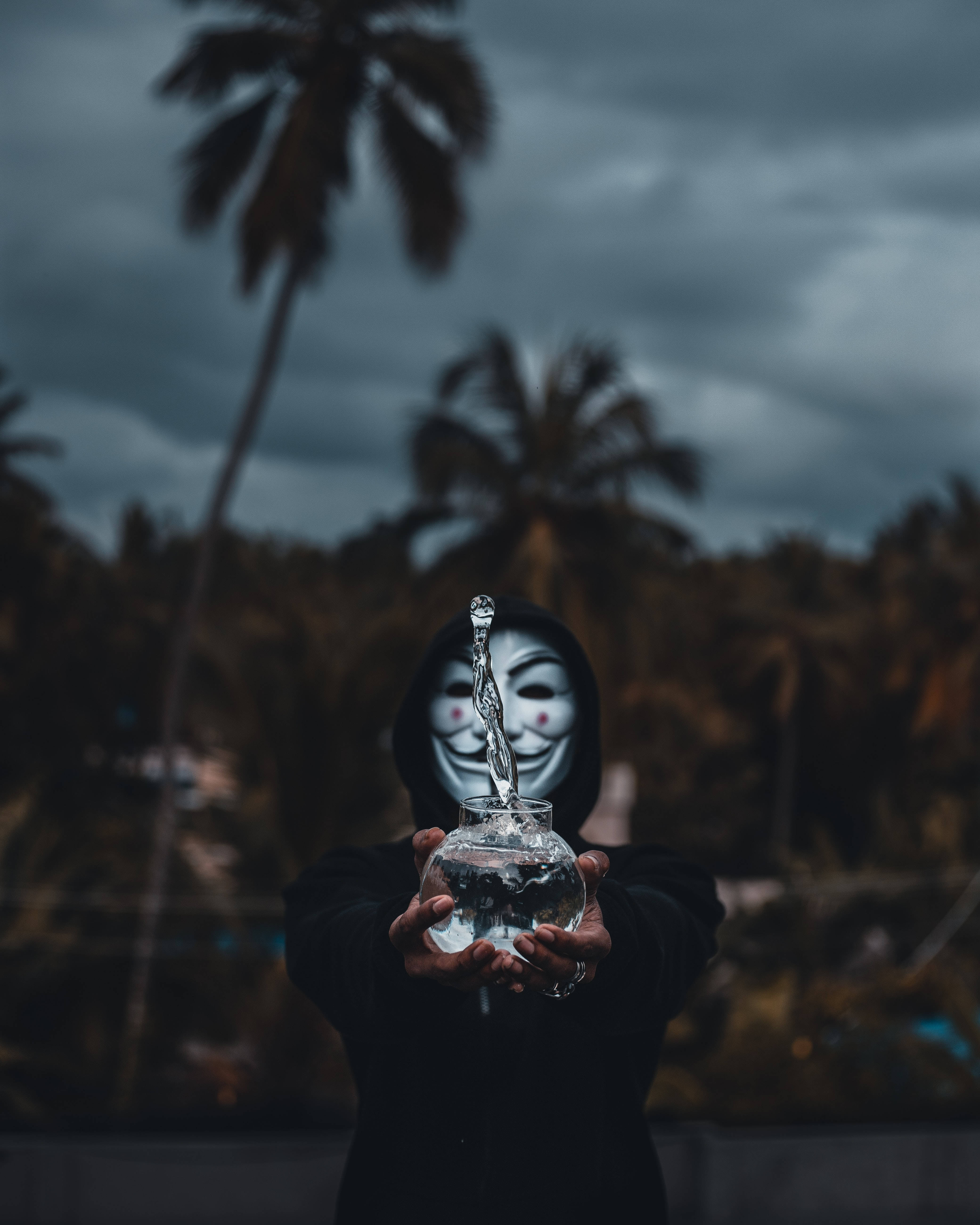 匿名 桌布 壁紙 — 一個穿著黑色連帽衫和匿名面具的人，在陰暗的棕櫚樹背景下，手持一個玻璃碗，水從勺子滴入其中。