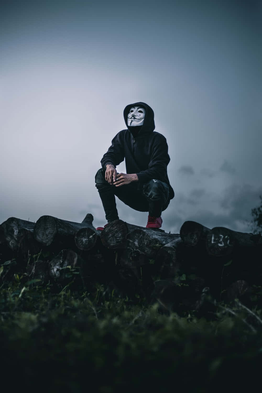 Mask 桌布 壁紙 — 一個穿著黑色連帽衫和標誌性白色面具的人蹲在一堆木頭上，天空陰沉多雲。