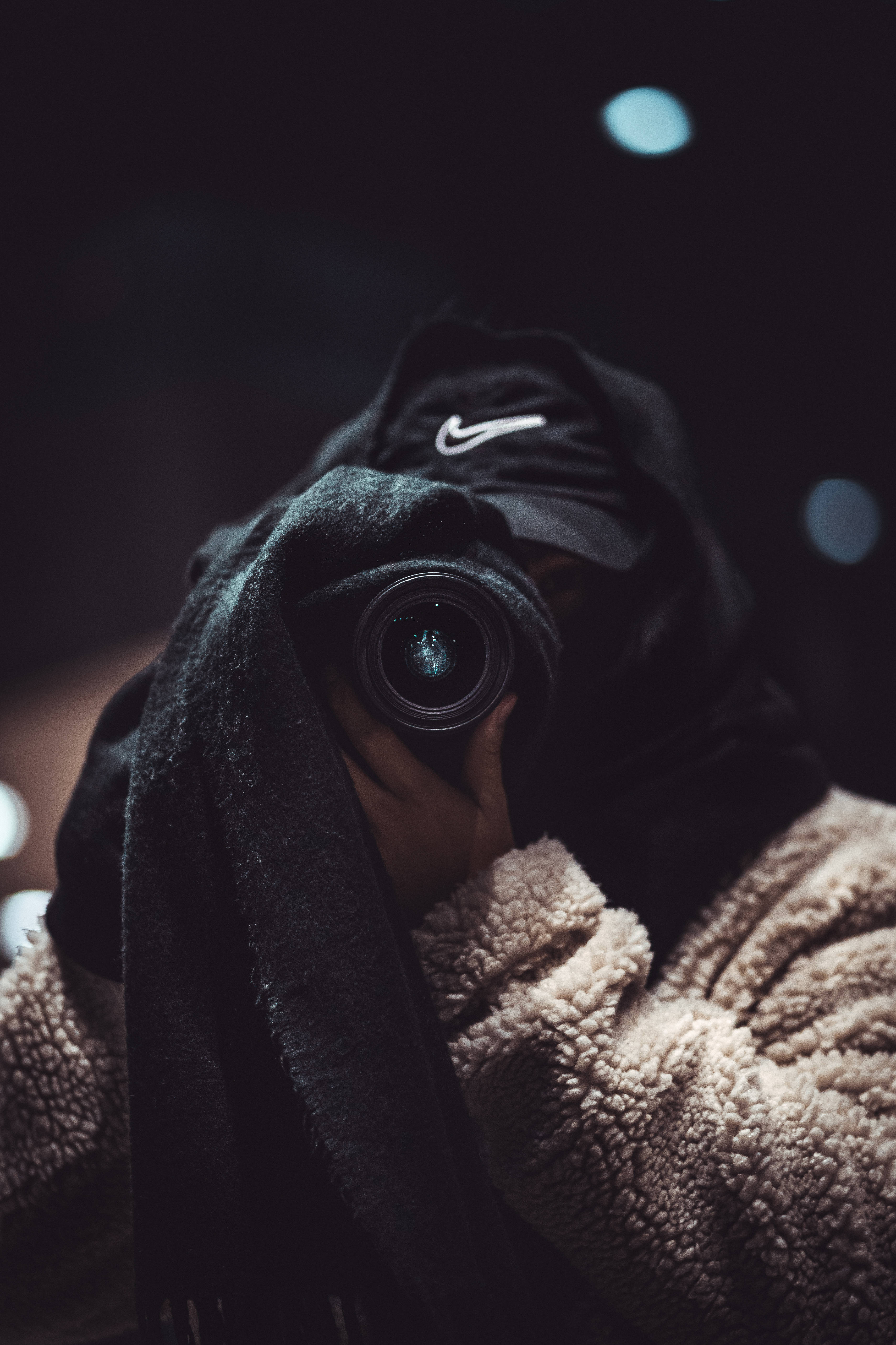 4K nike 桌布 壁紙 — 一個穿著黑色連帽衫和毛絨米色外套的人，在帶有柔和藍色散景燈光的黑暗背景下將相機舉到臉前。