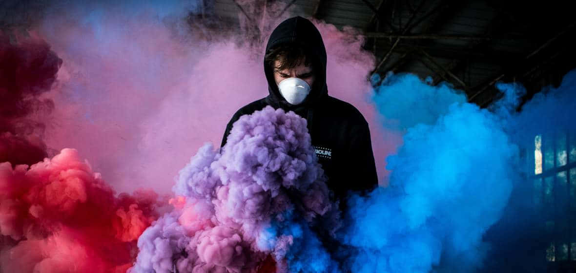 煙動圖 桌布 壁紙 — 一個穿著黑色連帽衫和白色口罩的人站在昏暗的工業空間中，周圍環繞著旋轉的紅色、粉紅色和藍色煙霧。