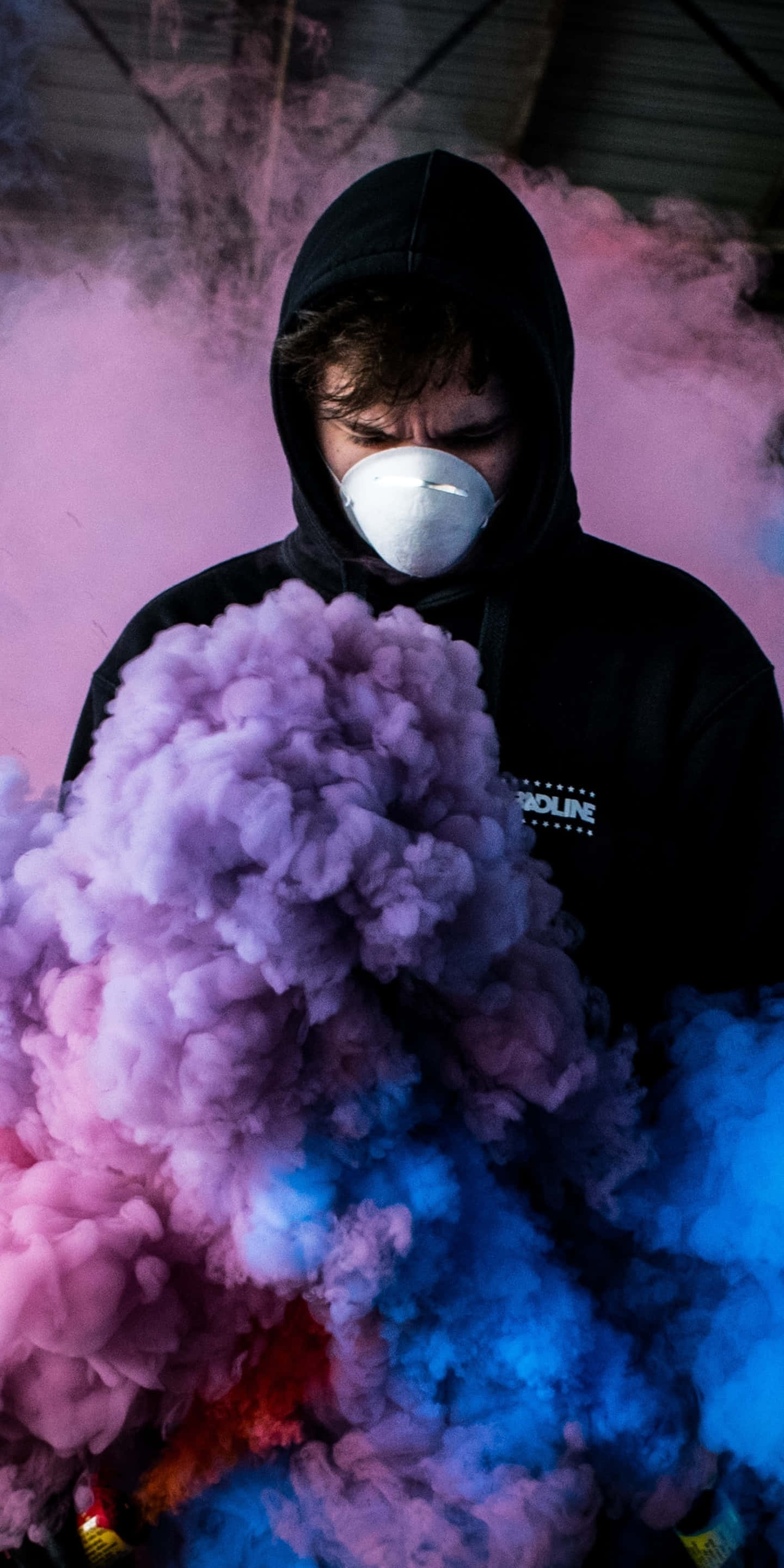 Mask 桌布 壁紙 — 一個穿著黑色連帽衫和白色口罩的人站在工業環境中，周圍環繞著旋轉的粉紅色和藍色煙霧。