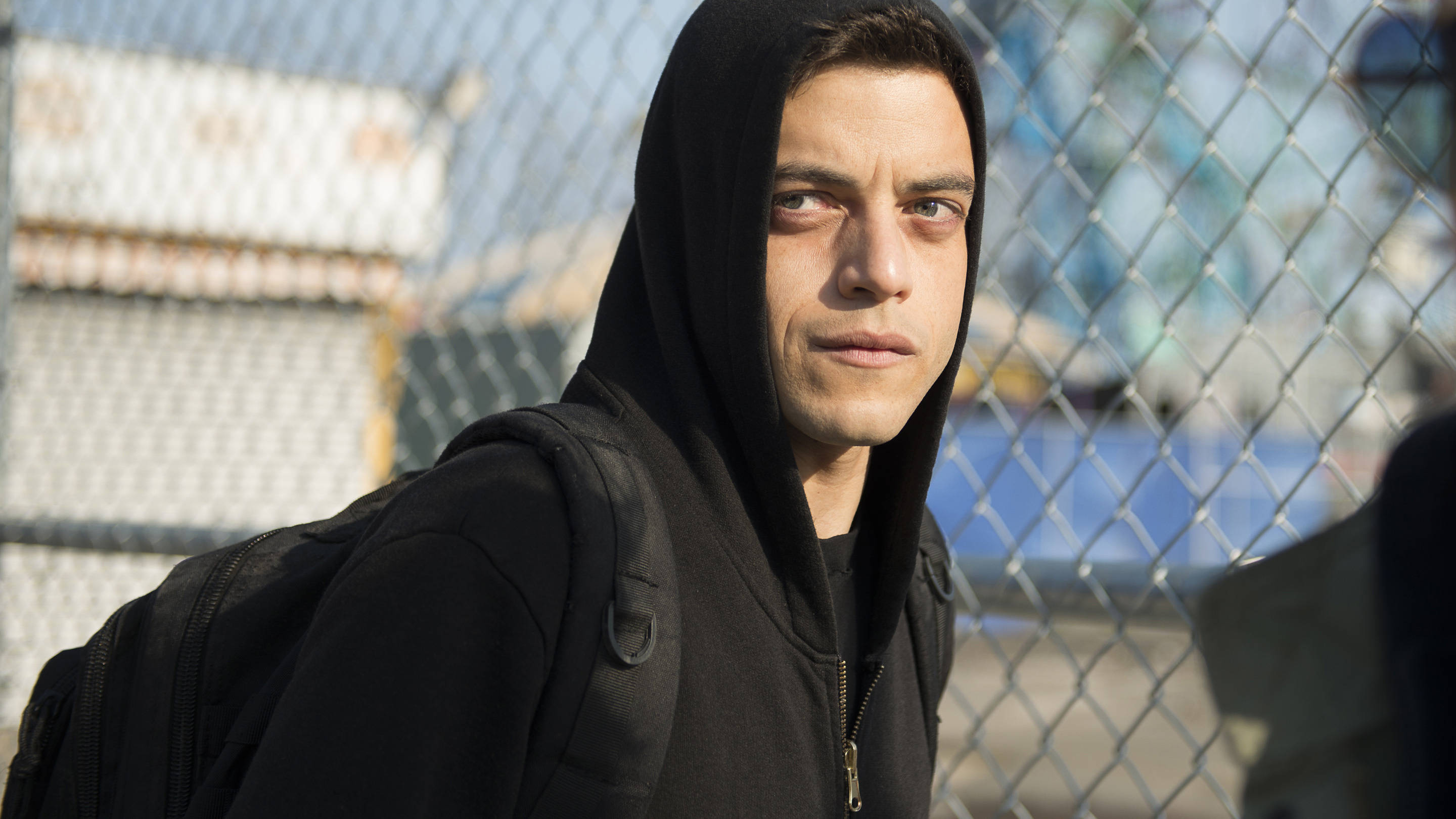 Mr Robot 桌布 壁紙 — 一個穿著黑色連帽衫和背包的年輕男子在明亮的日光下透過鐵絲網凝視。