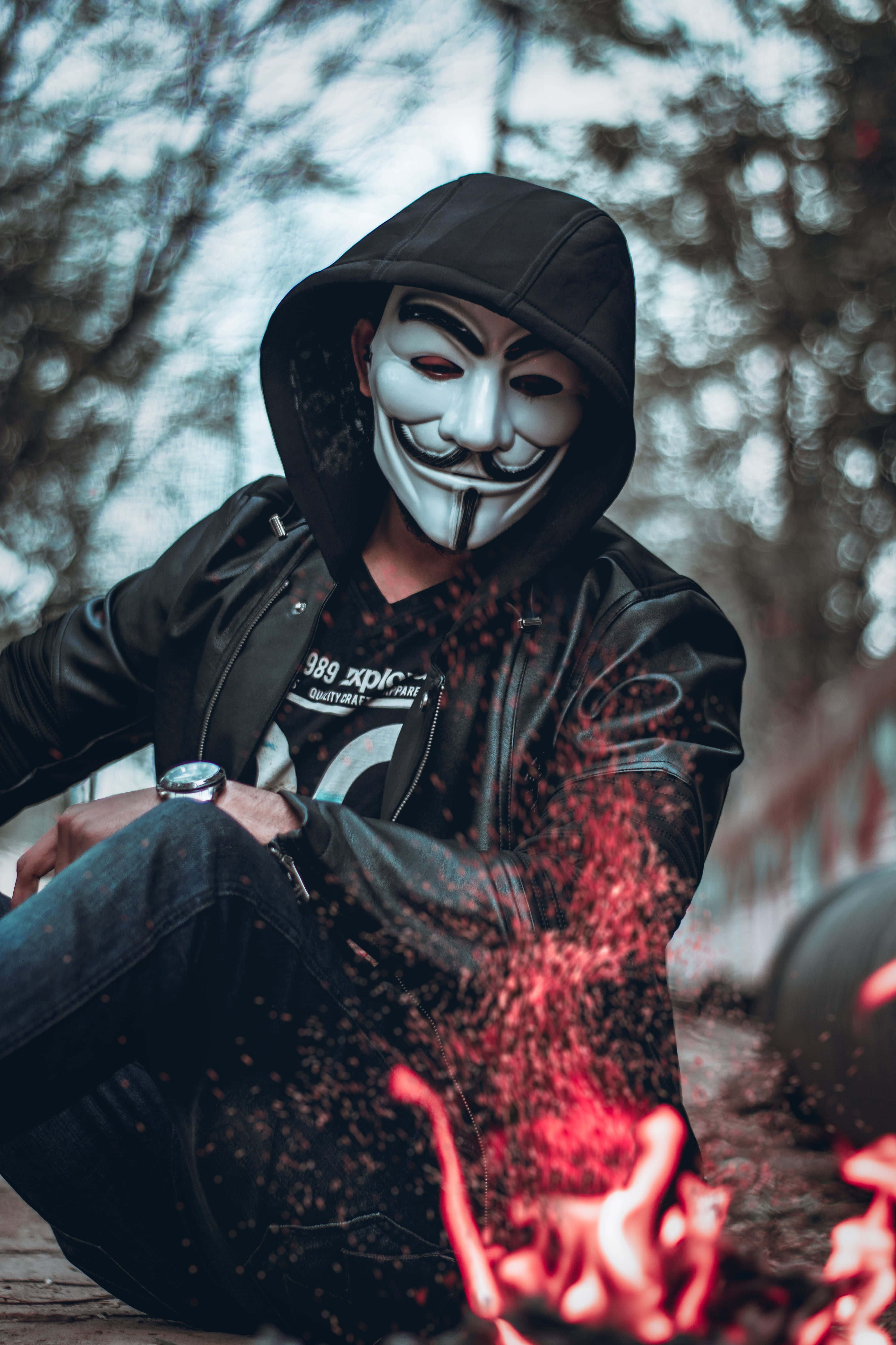 Embers 桌布 壁紙 — 一個穿著黑色連帽衫和蓋伊·福克斯面具的人坐在閃爍的紅色火堆旁，背景是模糊的樹木。