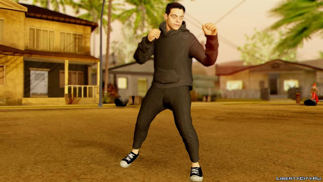 Rami Malek 桌布 壁紙 — 一個穿著黑色連帽衫和褲子的電子遊戲角色，在陽光明媚的郊區街道上擺出戰鬥姿勢，街道上有棕櫚樹和房屋。