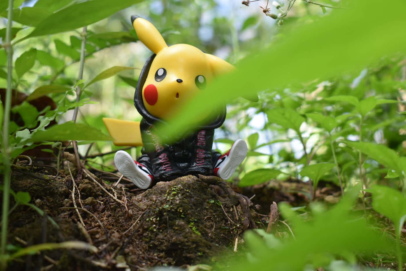 Pokemon Figures 桌布 壁紙 — 一個穿著黑色連帽衫和運動鞋的皮卡丘公仔坐在長滿青苔的石頭上，周圍是茂密的綠色樹葉。