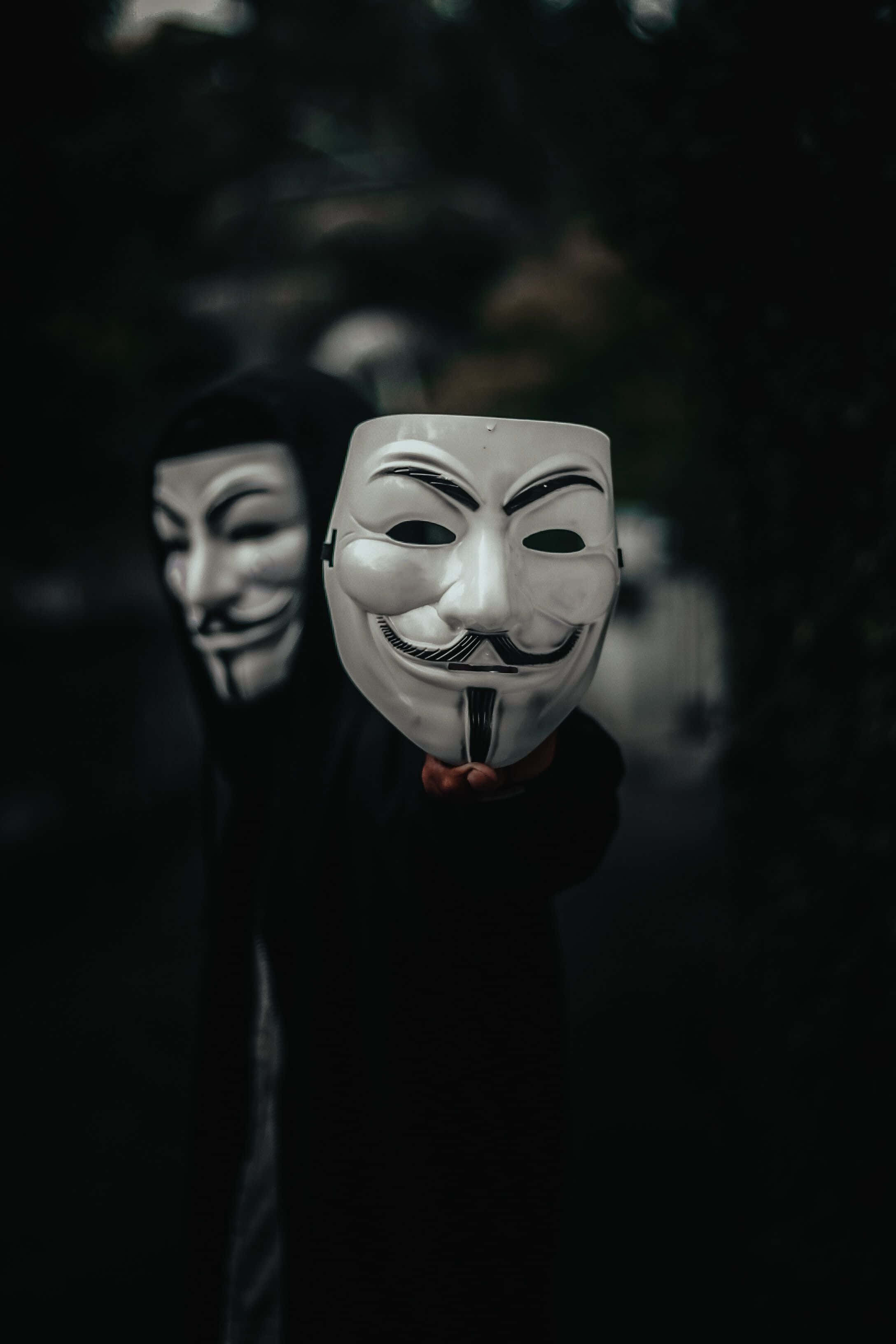 Mask Boy 桌布 壁紙 — 一個穿著黑色連帽衫的人，在黑暗模糊的背景前，將一個白色的蓋伊·福克斯面具向前舉起。