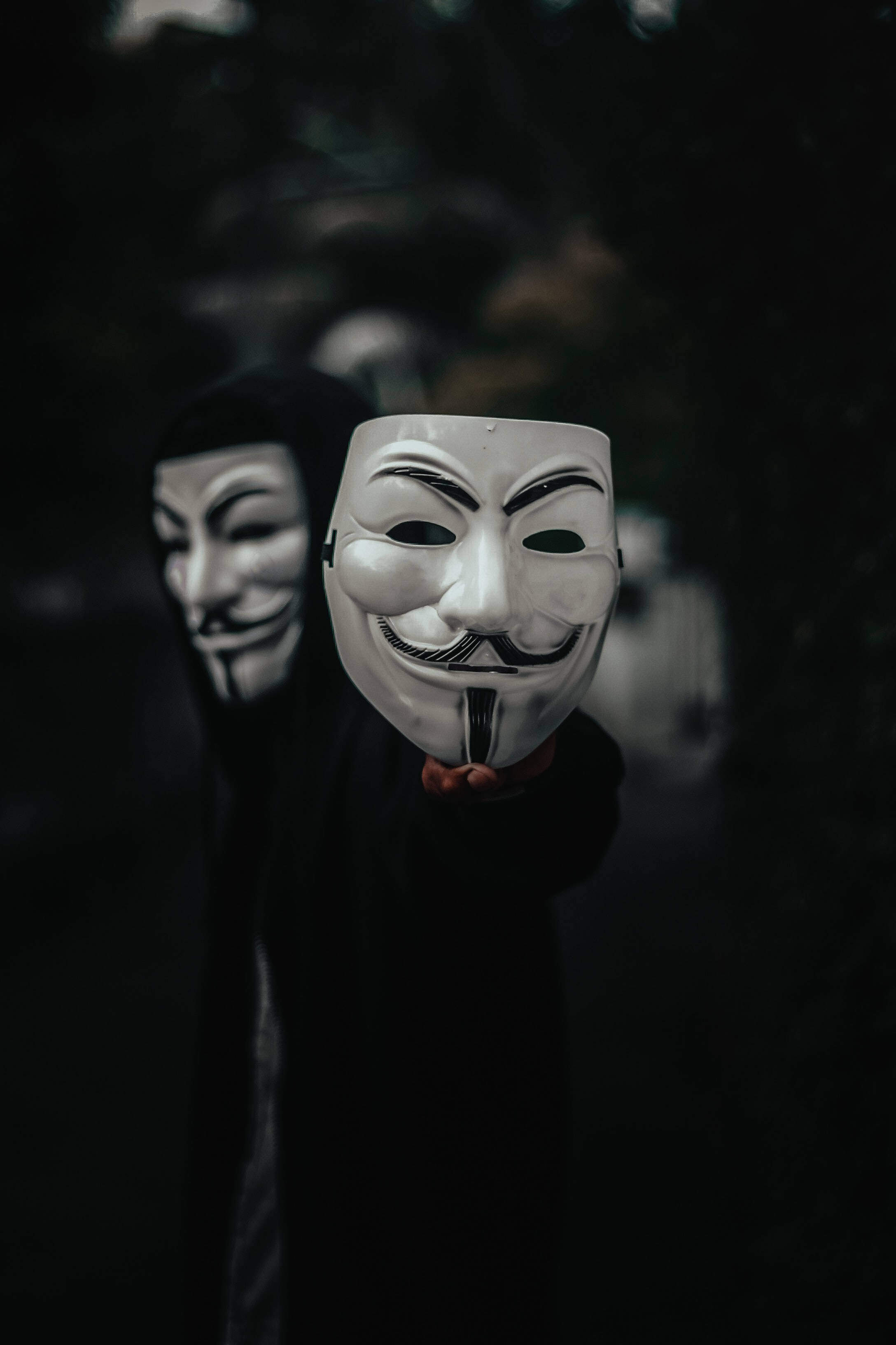 匿名 桌布 壁紙 — 一個穿著黑色連帽衫的人，在黑暗模糊的戶外環境中，將一個白色的蓋伊·福克斯面具向前舉起。