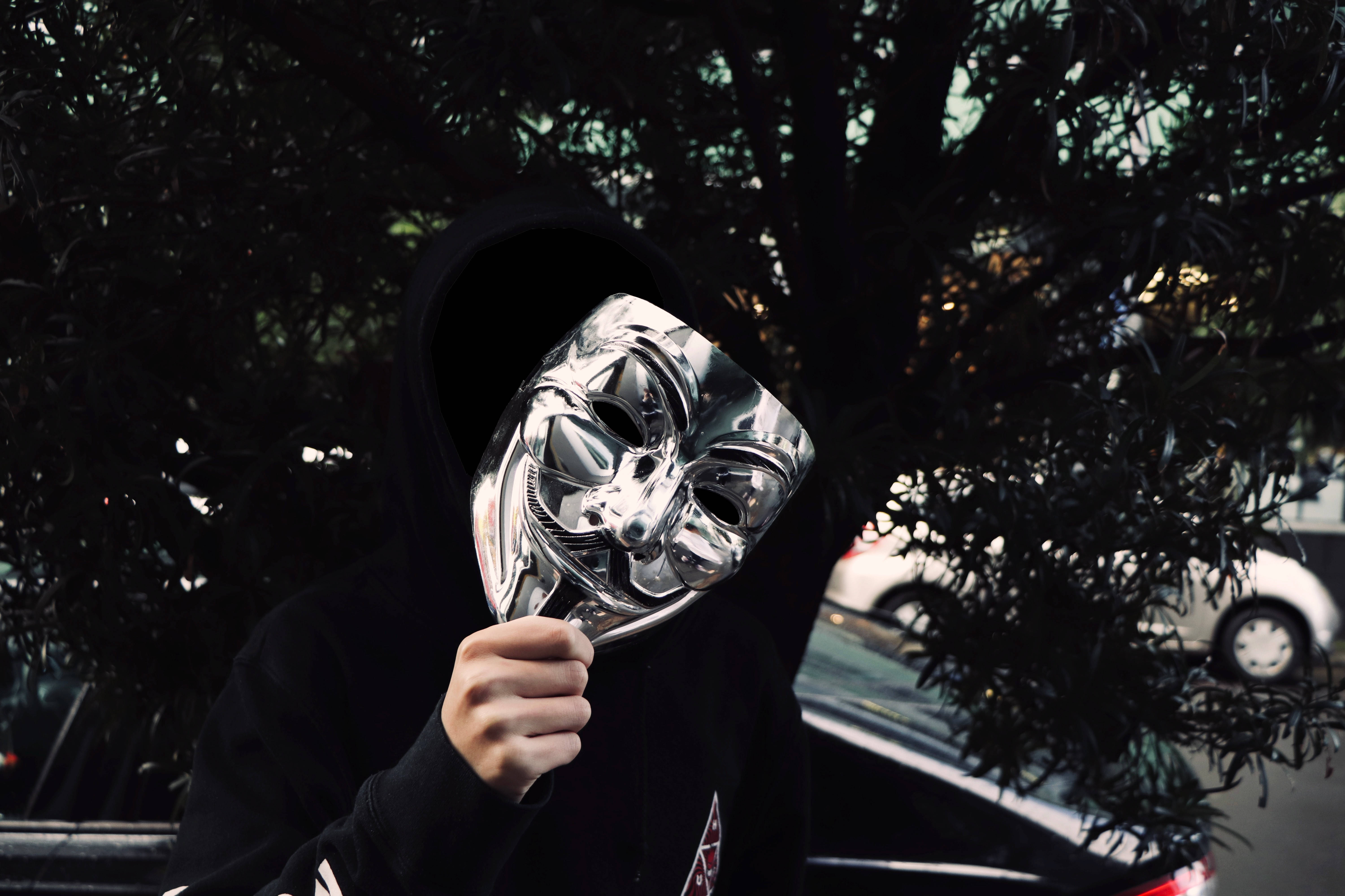 匿名 桌布 壁紙 — 一個穿著黑色連帽衫的人，在黑暗的樹葉中將閃亮的銀色蓋伊·福克斯面具舉在臉前。